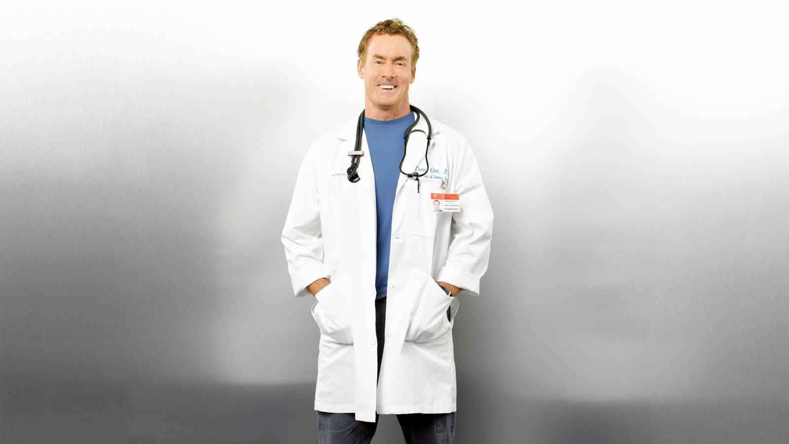 John C. Mcginley 桌布 壁紙 — 一位微笑的男醫生穿著白色實驗室外套，掛著聽診器，自信地站在柔和的灰色漸變背景前。