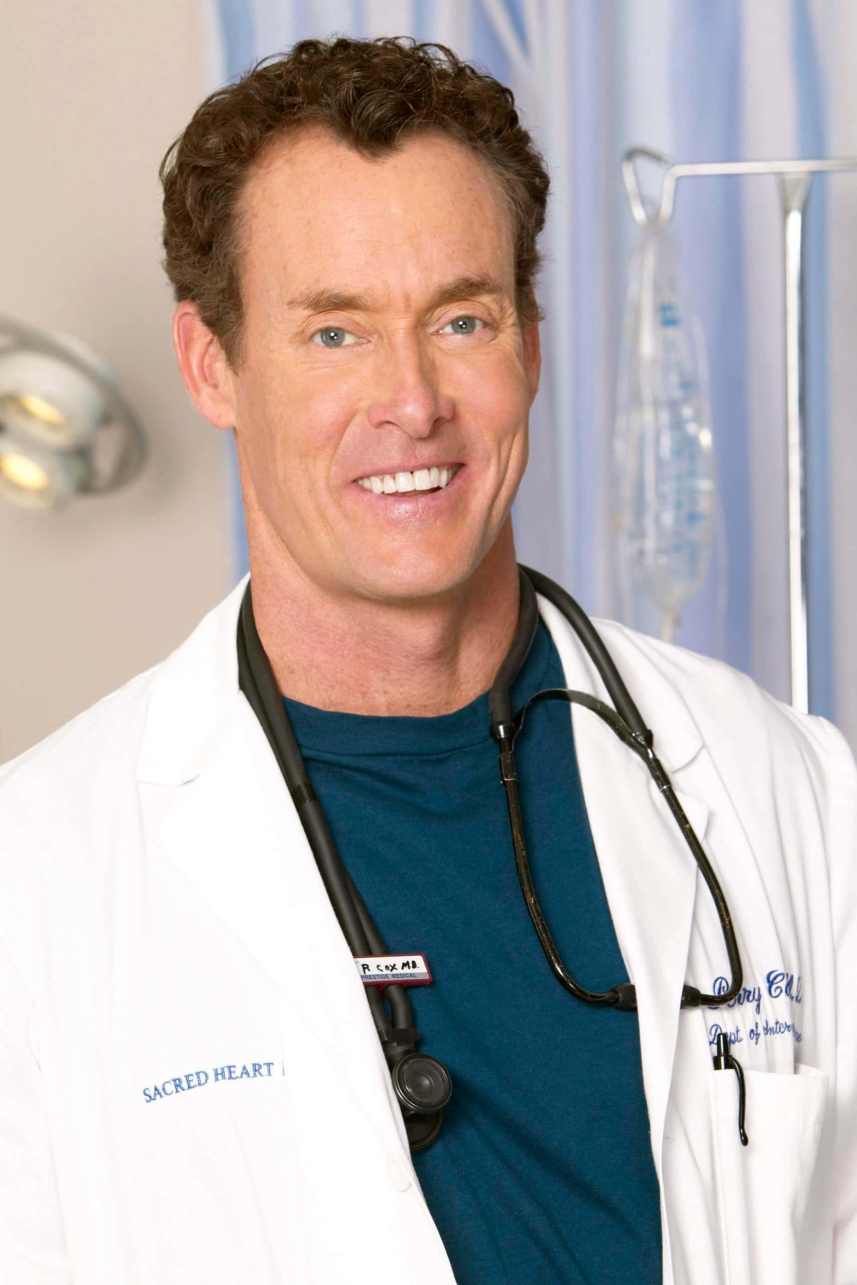 John C. Mcginley 桌布 壁紙 — 一位微笑的男醫生穿著白色實驗室外套，掛著聽診器，站在一間身後有醫療設備的醫院房間裏。