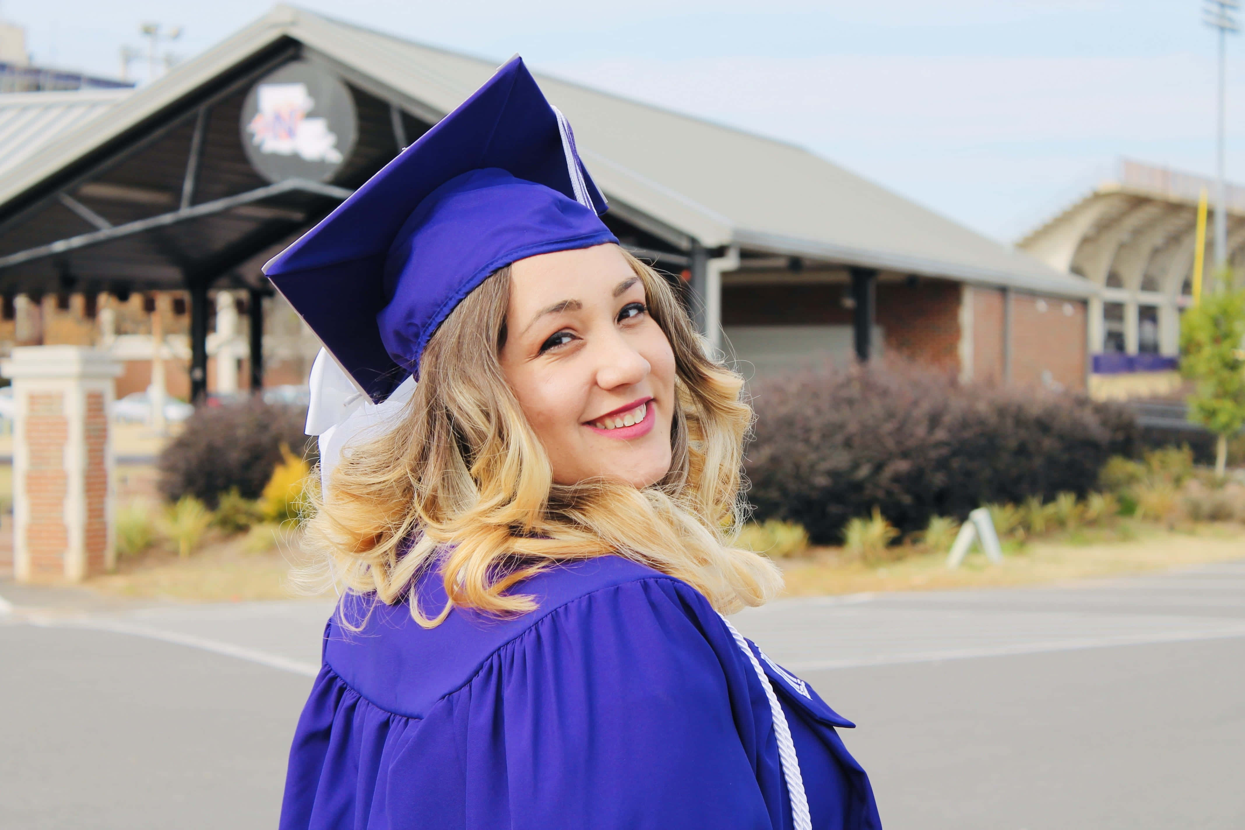 Graduation Ceremony 桌布 壁紙 — 一位微笑的畢業生身穿紫色畢業袍和四方帽，轉頭越過肩膀望向鏡頭。