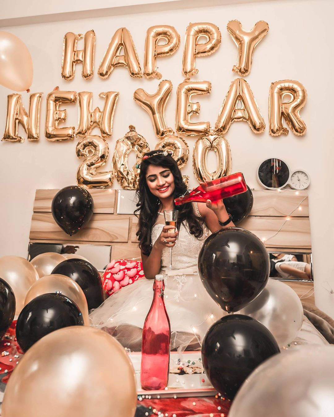 Nisha 桌布 壁紙 — 一位微笑的白裙女士在金色 'HAPPY NEW YEAR 2020' 氣球下倒香檳，周圍環繞著黑色和玫瑰金色氣球。