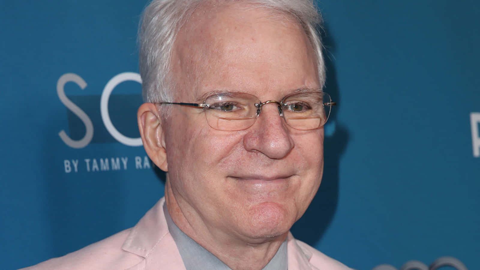 steve martin 桌布 壁紙 — 一位微笑的白髮戴眼鏡年長男士身穿粉紅色西裝，在藍色品牌背景前的特寫肖像。