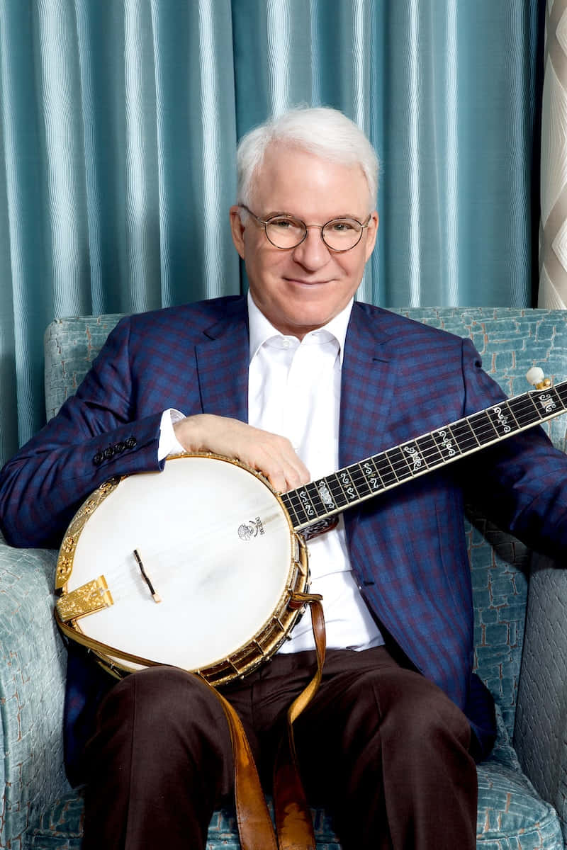 steve martin 桌布 壁紙 — 一位微笑的白髮戴眼鏡老翁，坐在青綠色扶手椅上，抱著班卓琴，背景是相配的窗簾。