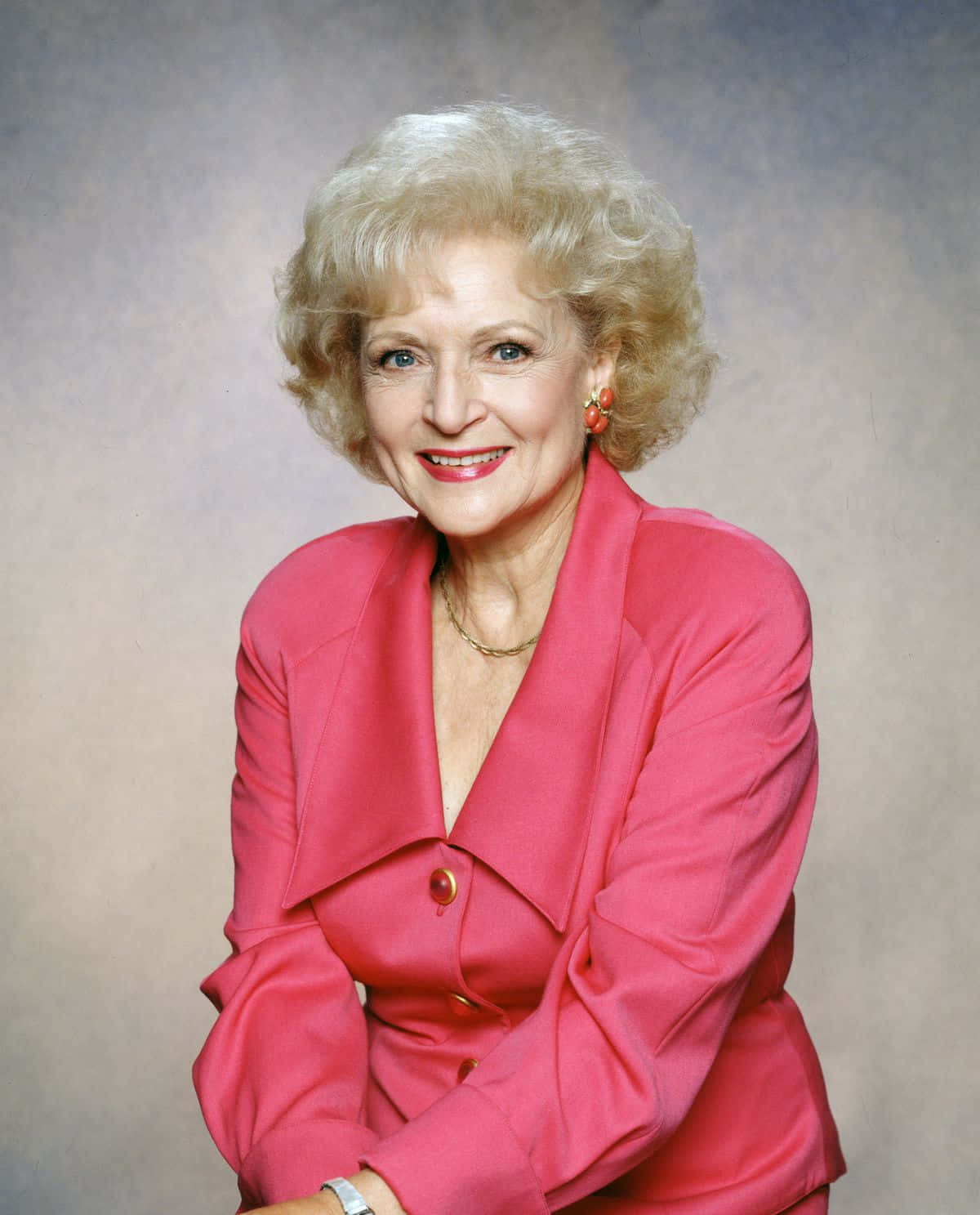 Betty White 桌布 壁紙 — 一位微笑的白髮捲曲年長女性，穿著鮮豔的粉紅色西裝外套，背景是柔和的灰色攝影棚。