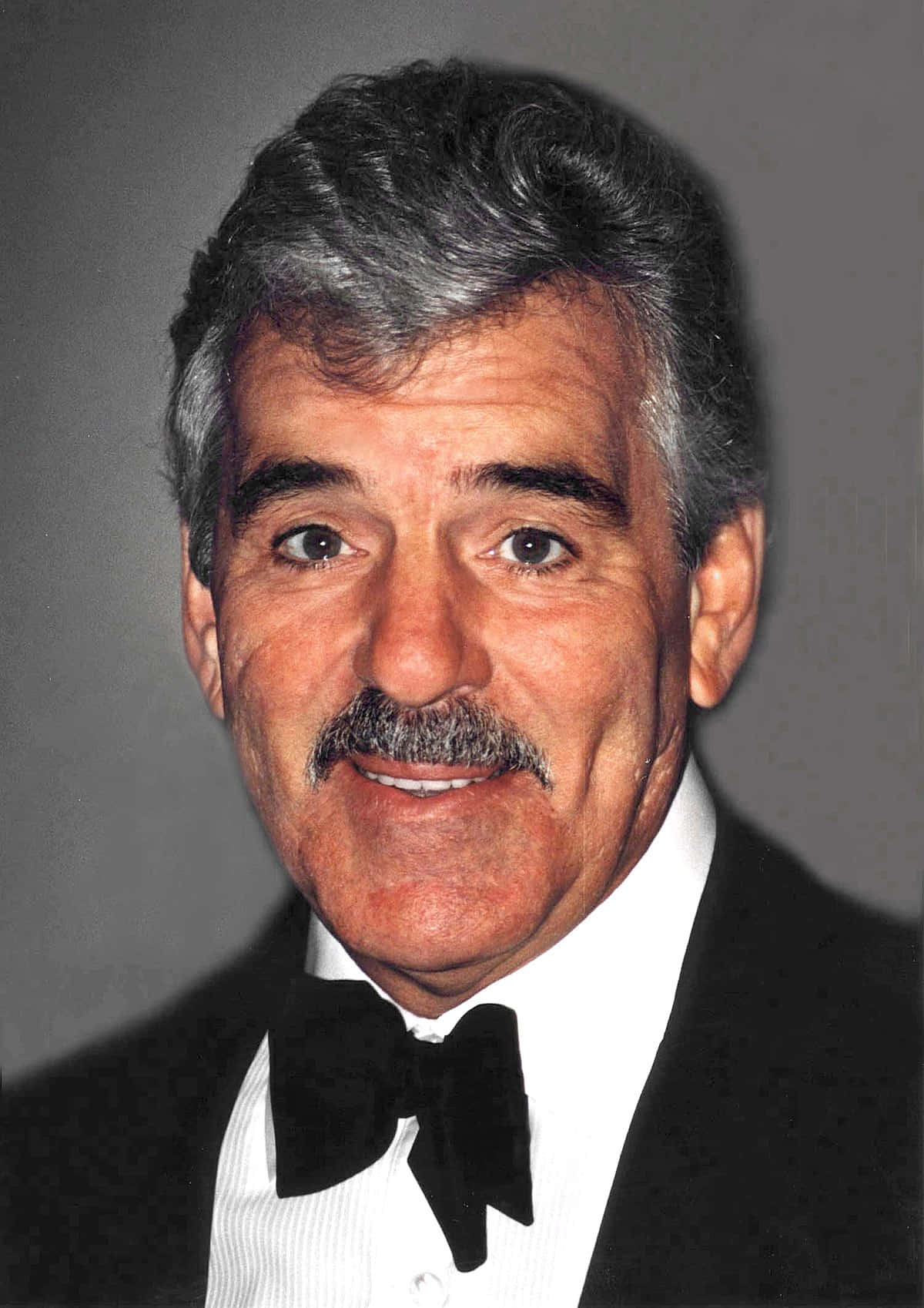 Dennis Farina 桌布 壁紙 — 一位微笑的白髮留鬍鬚男士，穿著黑色燕尾服和領結，背景為純灰色。