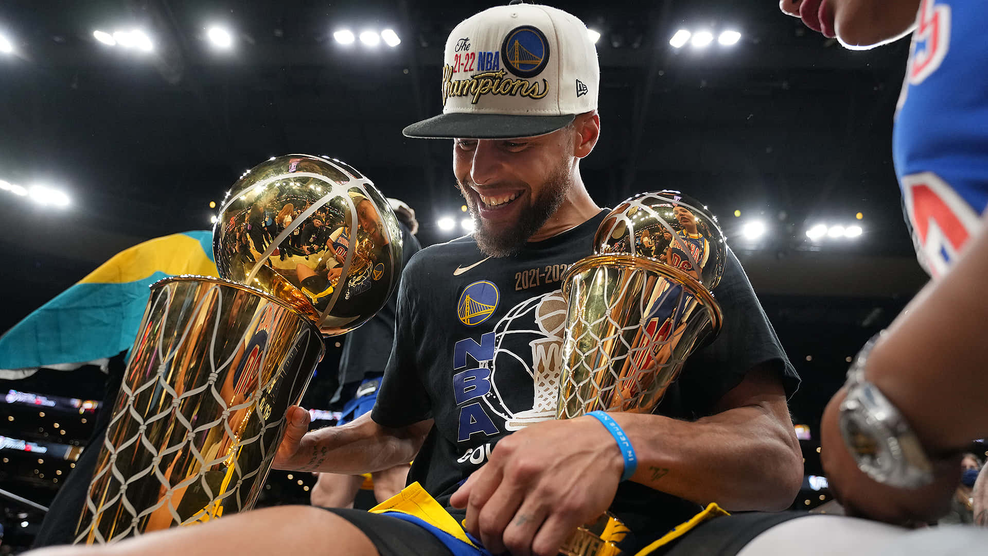 steph curry 桌布 壁紙 — 一位微笑的籃球運動員戴著冠軍帽，在場館燈光下手持兩個閃亮的NBA獎盃。
