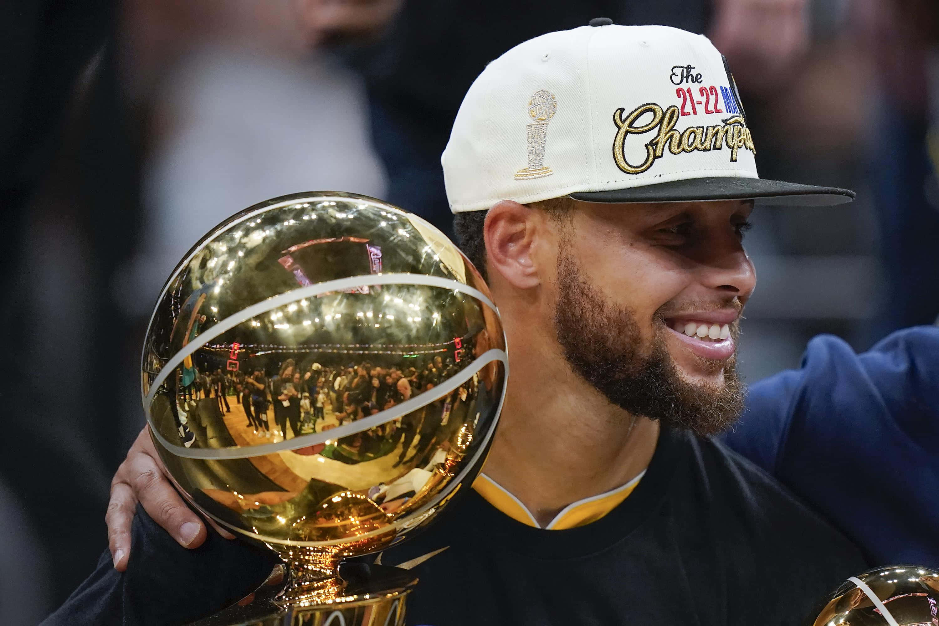 史提芬·居里 桌布 壁紙 — 一位微笑的籃球運動員自豪地舉起閃亮的NBA冠軍獎盃，同時戴著'21-22 Champion'帽子。