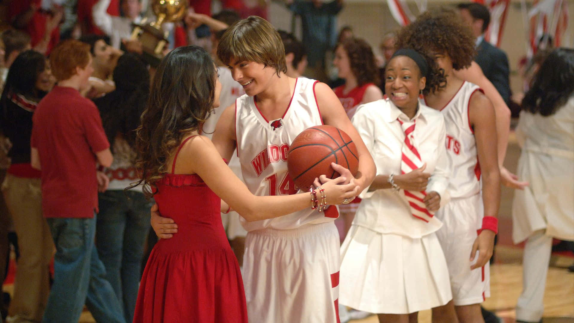 High School Musical 桌布 壁紙 — 一位微笑的籃球運動員身穿白色球衣，在體育館球場上抱著一個球，同時擁抱著一位穿紅色連衣裙的女孩。