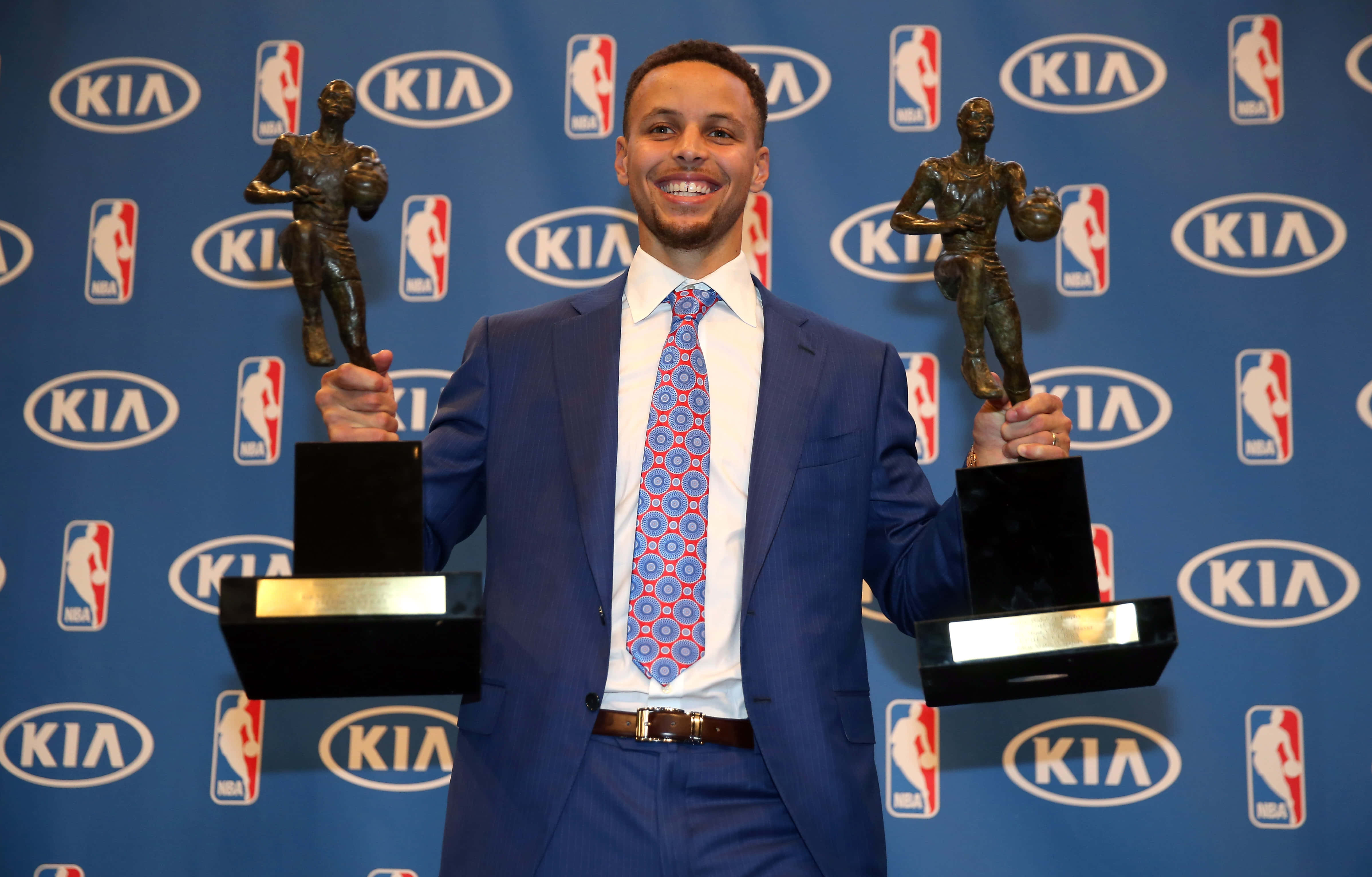 steph curry 桌布 壁紙 — 一位微笑的籃球運動員身穿藍色西裝，在帶有NBA和Kia標誌的藍色背景前手持兩個獎盃。