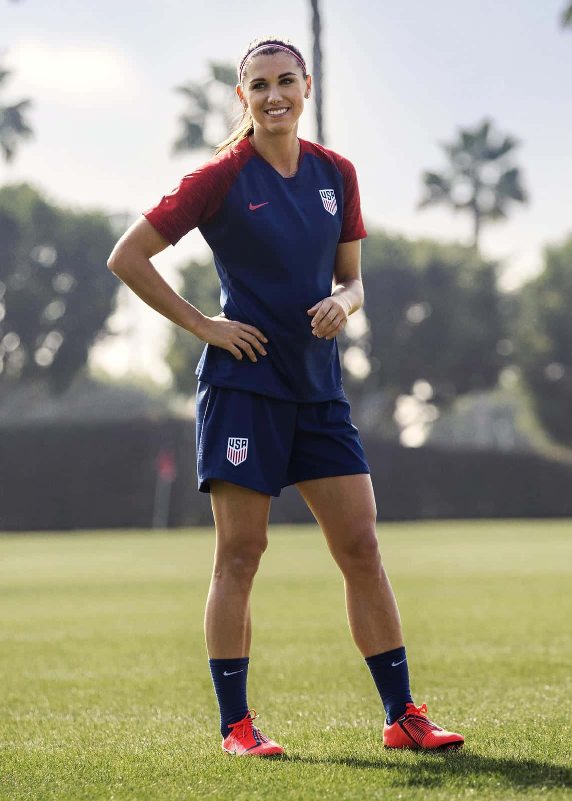 Alex Morgan 桌布 壁紙 — 一位微笑的美國國家隊女足球員，自信地站在陽光明媚的草地上，背景有棕櫚樹。