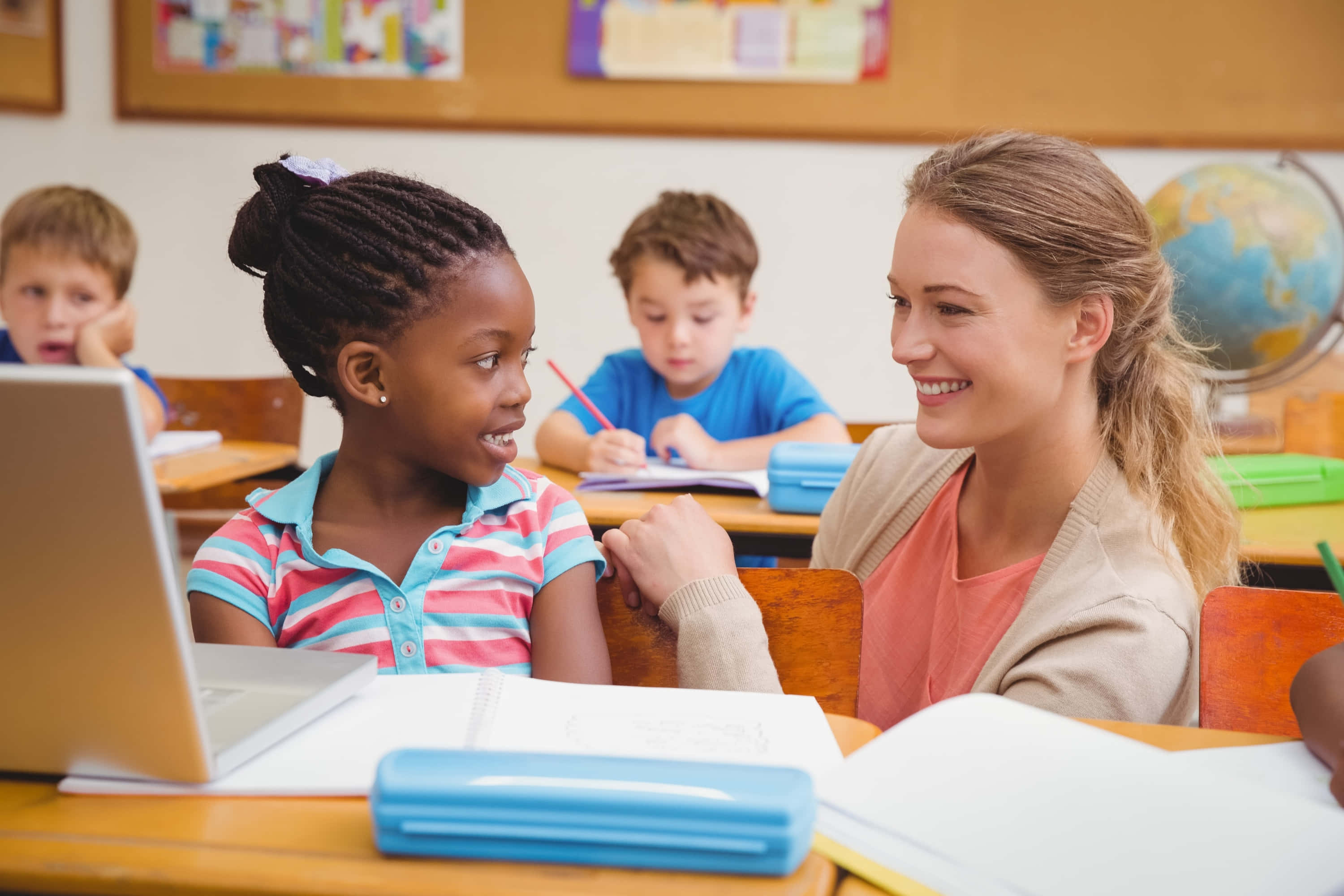 Learning 桌布 壁紙 — 一位微笑的老師在課室中與一位小女孩互動，其他學生在書桌上學習，背景有一個地球儀。