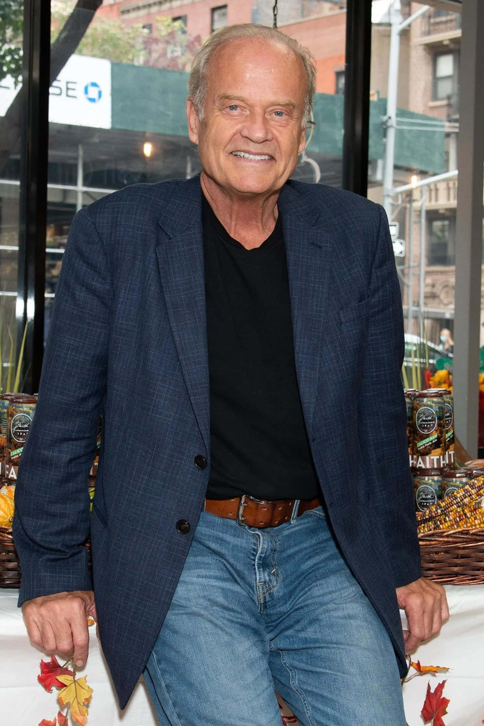 Kelsey Grammer 桌布 壁紙 — 一位微笑的老年男士穿著藍色格紋西裝外套和牛仔褲，在室內擺姿勢，附近有秋葉和罐頭食品。