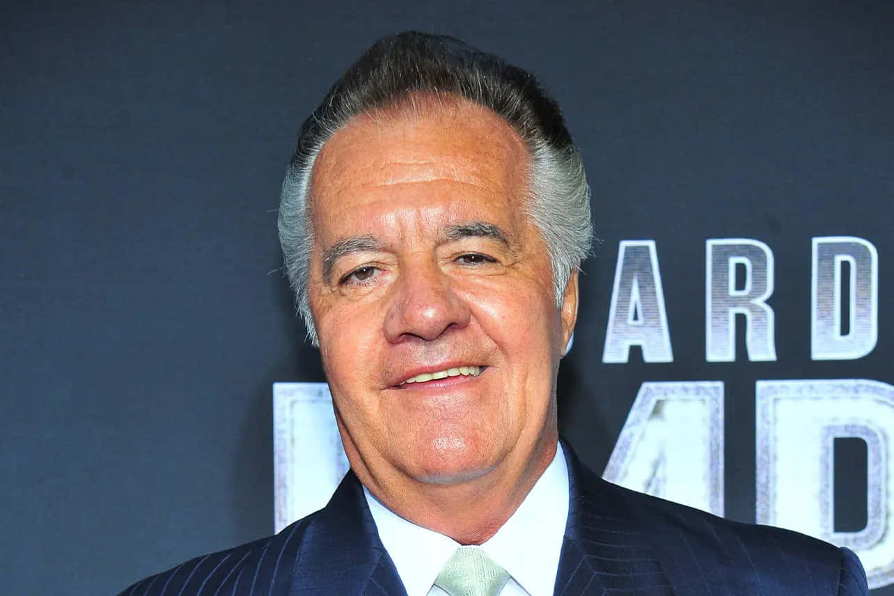 Tony Sirico 桌布 壁紙 — 一位微笑的老年男士身穿深色細條紋西裝和淺綠色領帶，在帶有部分銀色文字的深色背景前擺姿勢。