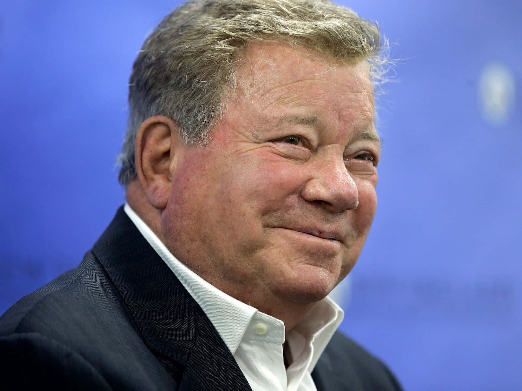 william shatner 桌布 壁紙 — 一位微笑的老年男士身穿深色西裝，以純藍色背景為襯的 close-up 肖像。