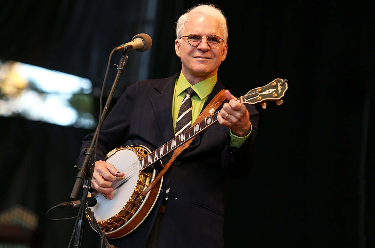 steve martin 桌布 壁紙 — 一位微笑的老年男子身穿西裝，在明亮的燈光下於舞台上彈奏班卓琴，旁邊有麥克風。
