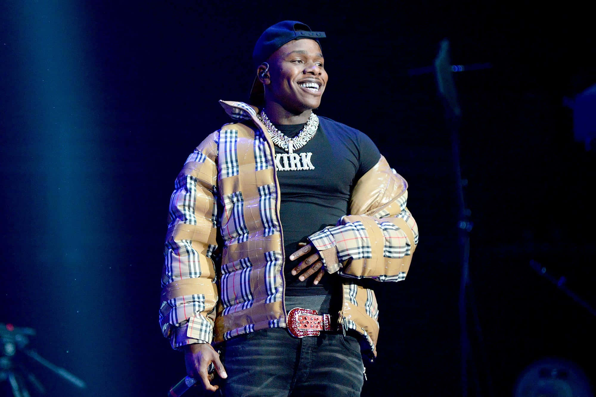 Dababy 桌布 壁紙 — 一位微笑的表演者穿著格仔羽絨外套和帽子，站在藍色燈光下的黑暗舞台上。
