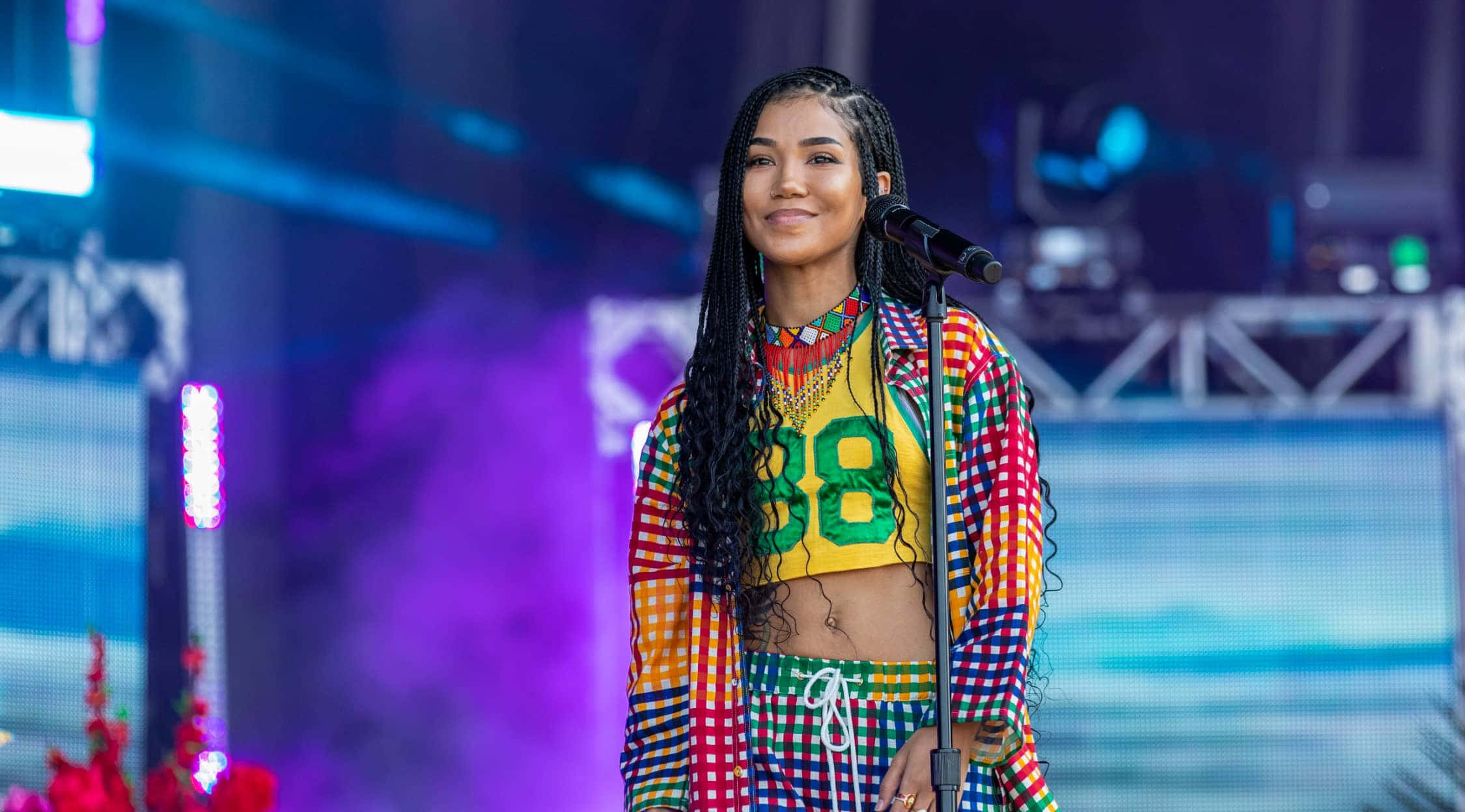 jhene aiko 桌布 壁紙 — 一位微笑的表演者穿著色彩繽紛的格仔服裝，在閃爍燈光下自信地站在充滿活力的舞台上。