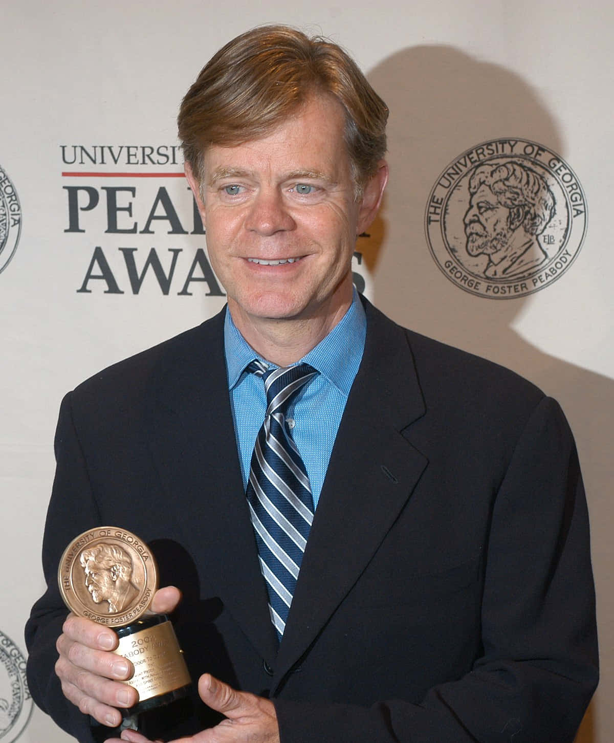 william h. macy 桌布 壁紙 — 一位微笑的西裝男子在品牌背景前手持皮博迪獎獎盃。