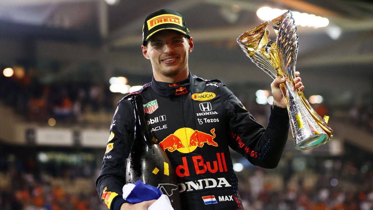 Verstappen 桌布 壁紙 — 一位微笑的賽車手身穿Red Bull賽車服，在明亮的體育場燈光下手持閃亮的獎盃和香檳瓶。