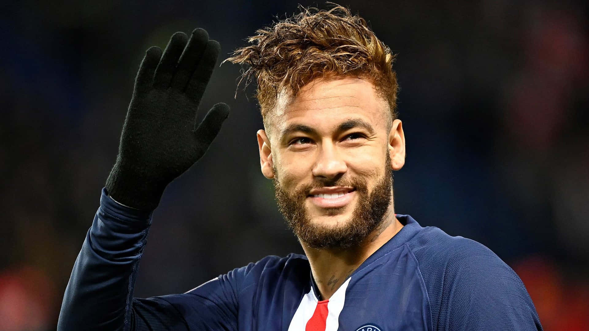 Neymar 桌布 壁紙 — 一位微笑的足球員，頭髮蓬亂，留著鬍鬚，穿著藍色球衣和黑色手套，熱情地向球迷揮手。