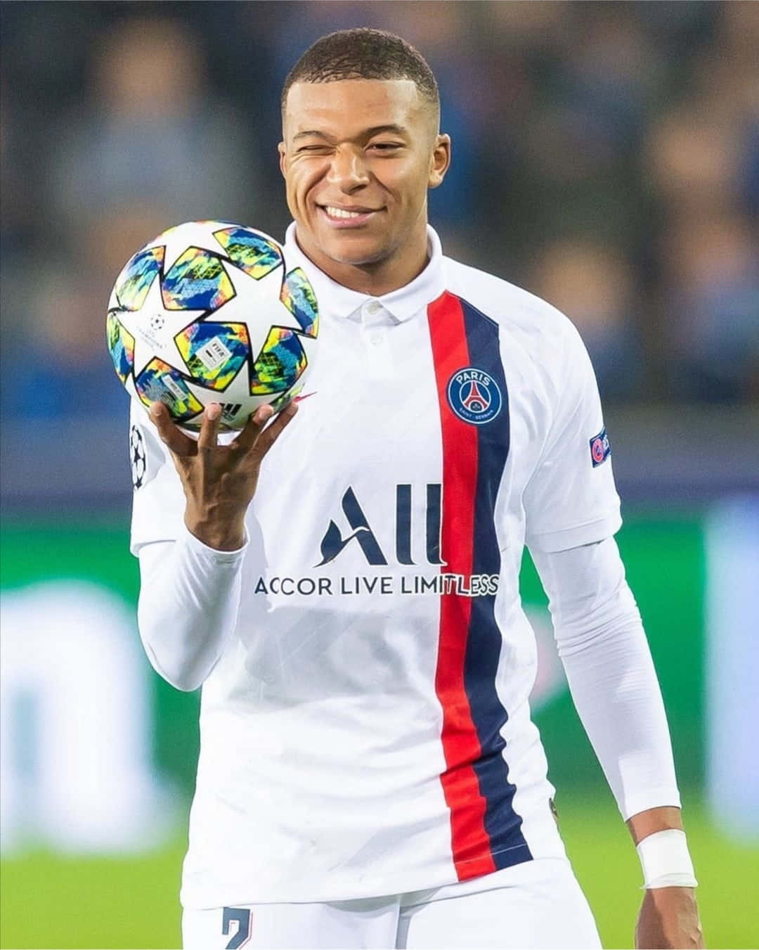 Mbappe 桌布 壁紙 — 一位微笑的足球員身穿巴黎聖日耳曼球衣，在球場上拿著色彩繽紛的歐聯足球。
