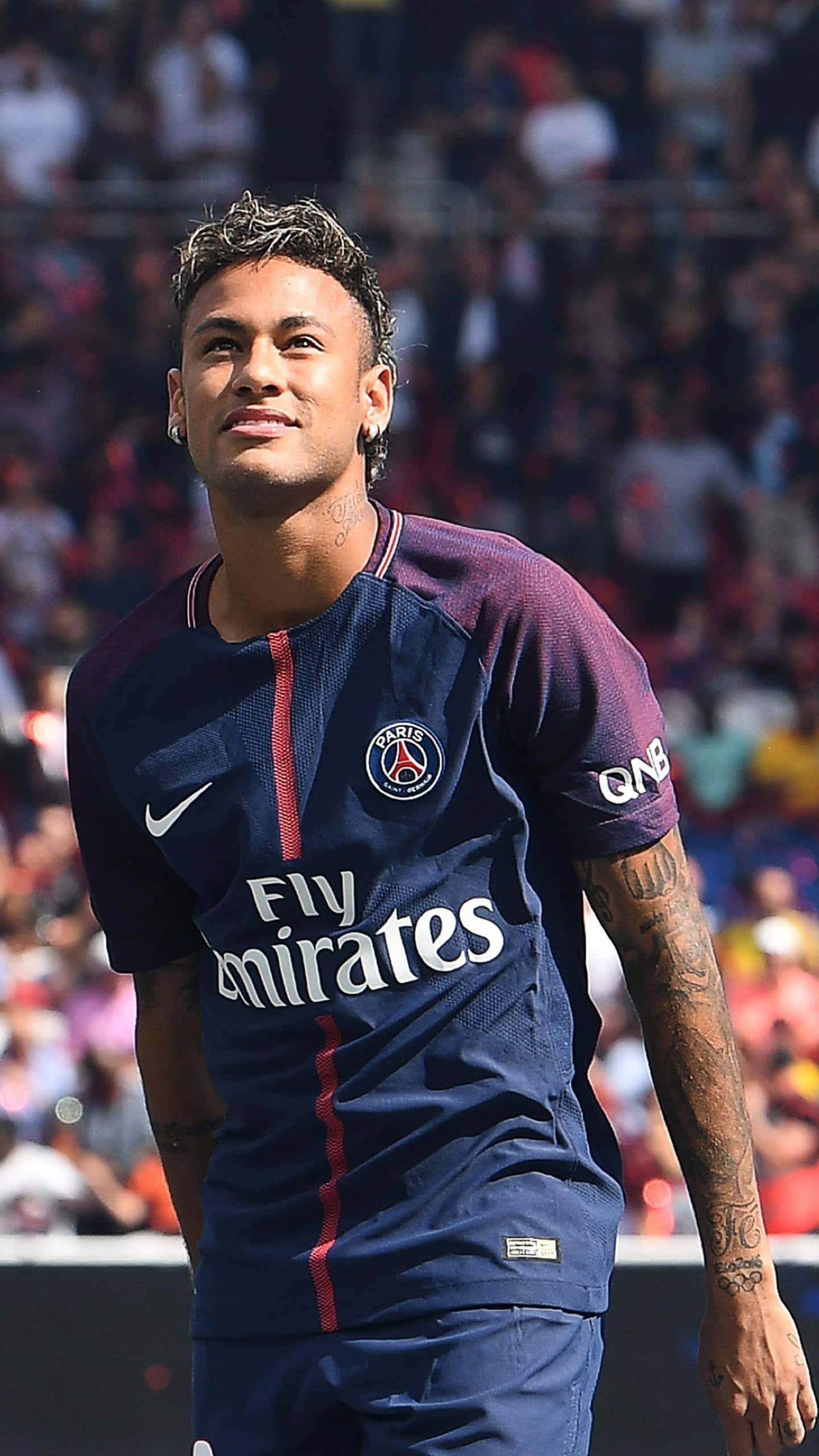 Neymar 桌布 壁紙 — 一位微笑的足球員身穿巴黎聖日耳曼球衣，自信地站在陽光普照的球場上，身後是模糊的人群。
