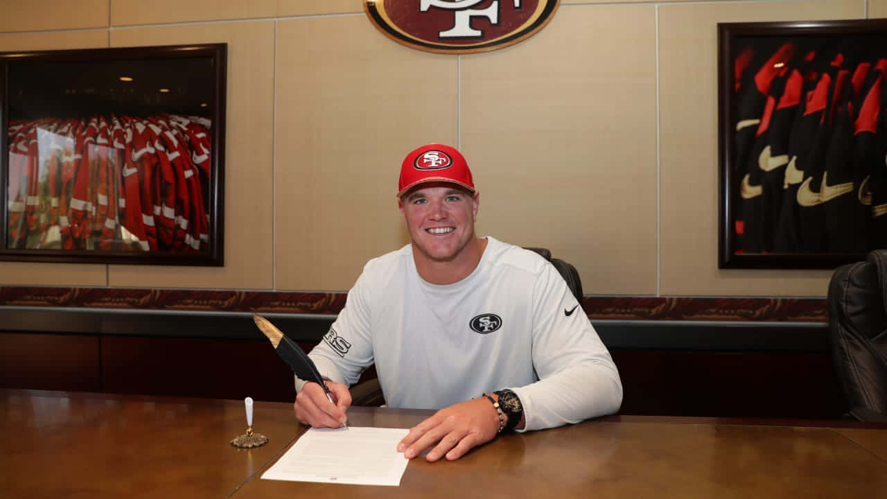 Mike Mcglinchey 桌布 壁紙 — 一位微笑的足球運動員，戴著紅色帽子，身穿白色球衣，在球隊辦公室的木桌上簽署合約。