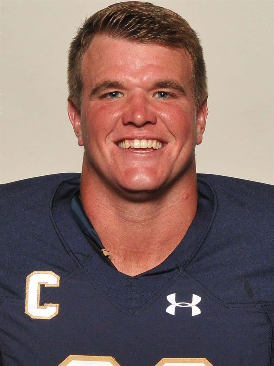 Mike Mcglinchey 桌布 壁紙 — 一位微笑的足球運動員，身穿海軍藍球衣，帶有金色字母和隊長 'C' 徽章。