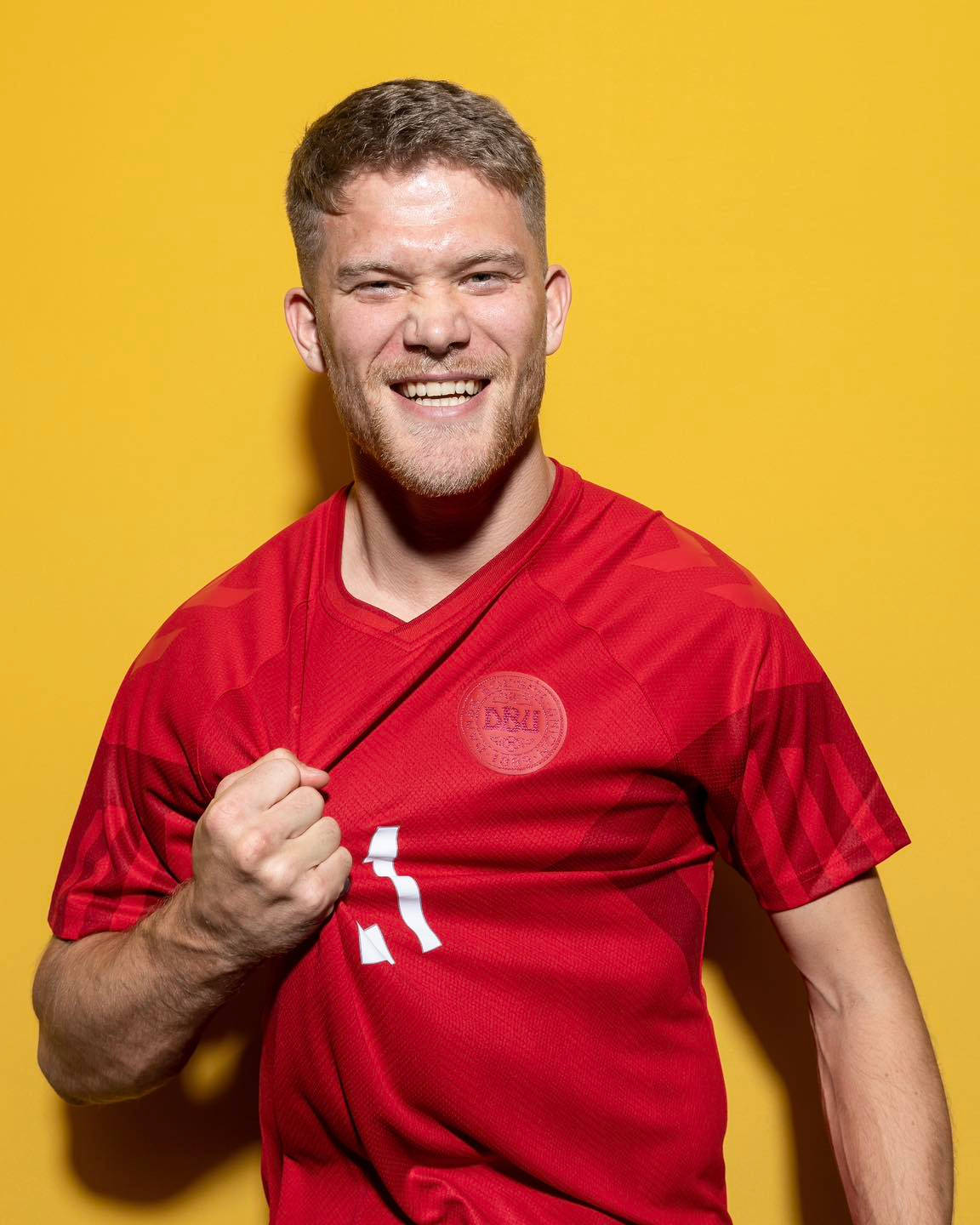 Football Players 桌布 壁紙 — 一位微笑的足球運動員身穿紅色球衣，在充滿活力的黃色背景前自信地擺姿勢。
