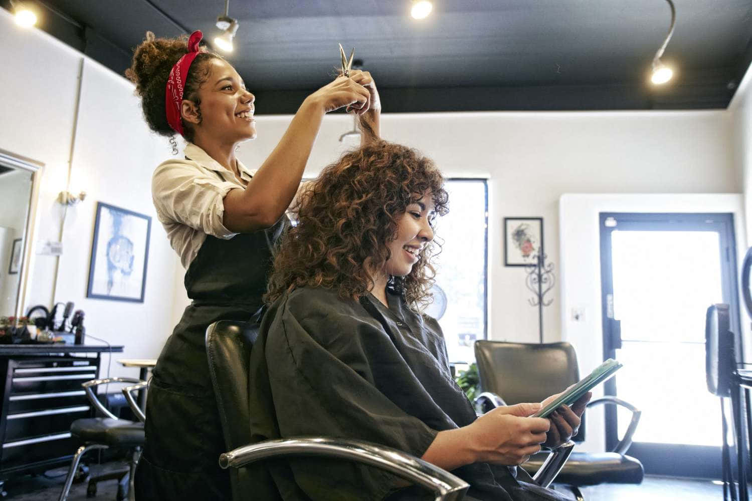 Hair Salon 桌布 壁紙 — 一位微笑的造型師在明亮現代的美髮沙龍中為正在閱讀雜誌的客戶修剪捲髮。