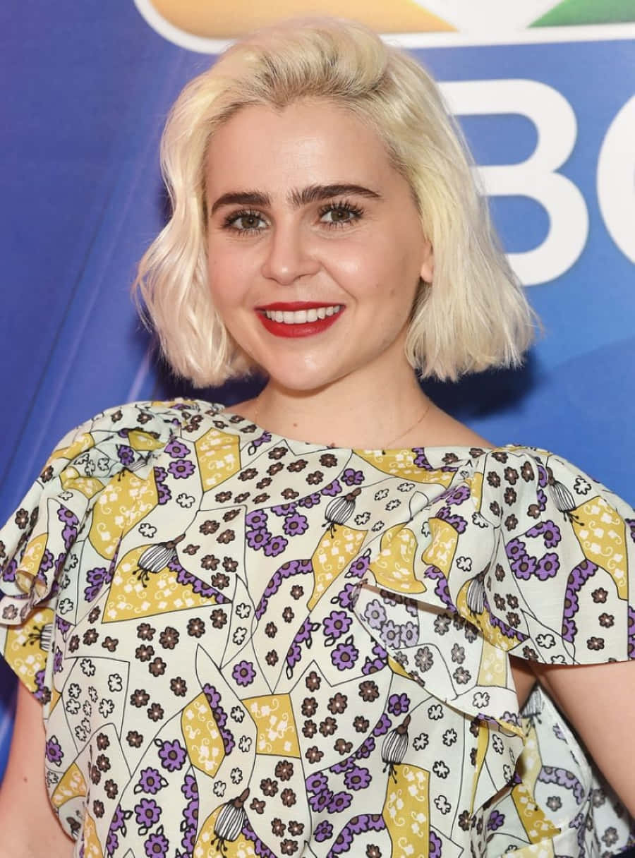 Mae Whitman 桌布 壁紙 — 一位微笑的金色波波頭女性，塗著紅色唇膏，穿著黃紫色花卉圖案襯衫，背景為藍色品牌背景。