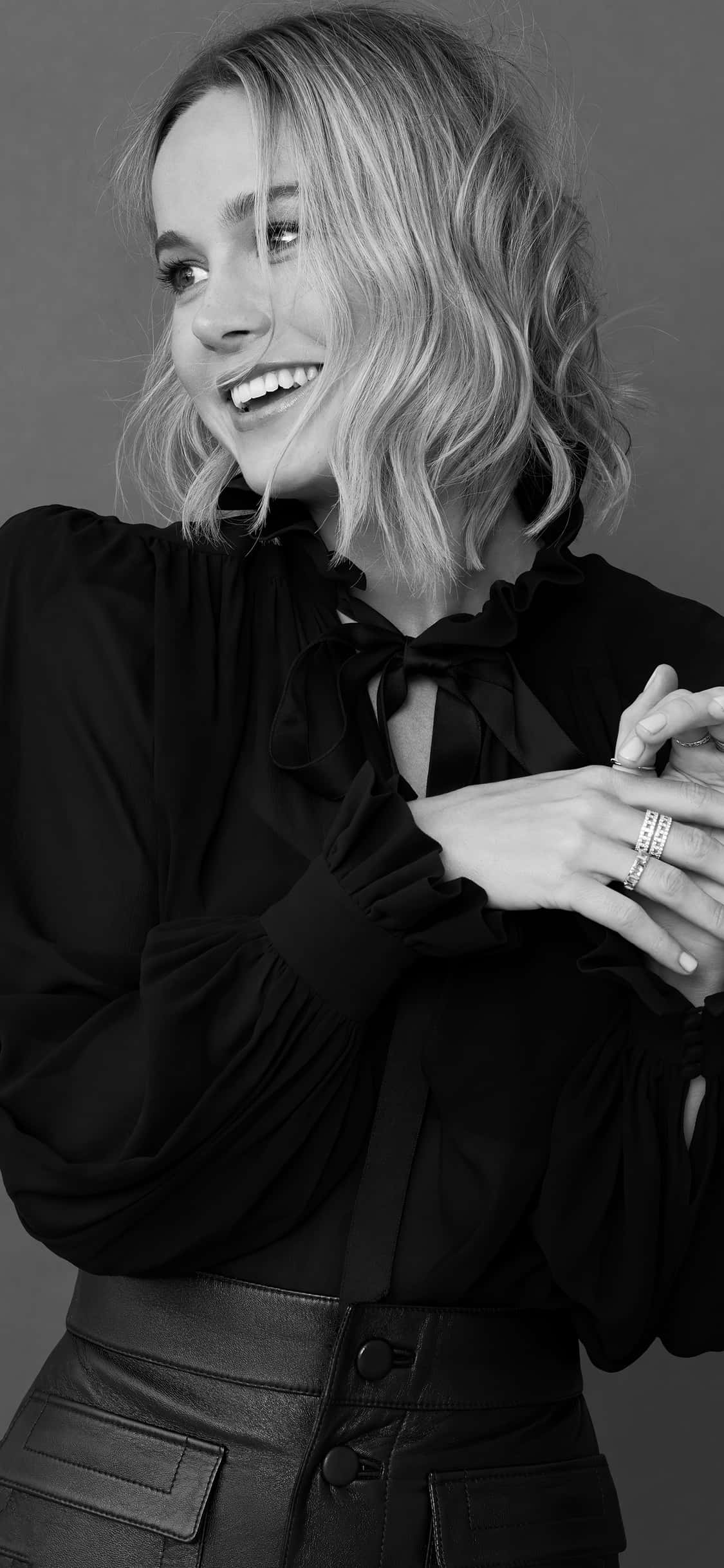 brie larson 桌布 壁紙 — 一位微笑的金色波浪長髮女性，在黑白工作室肖像中穿著黑色襯衫和皮褲。