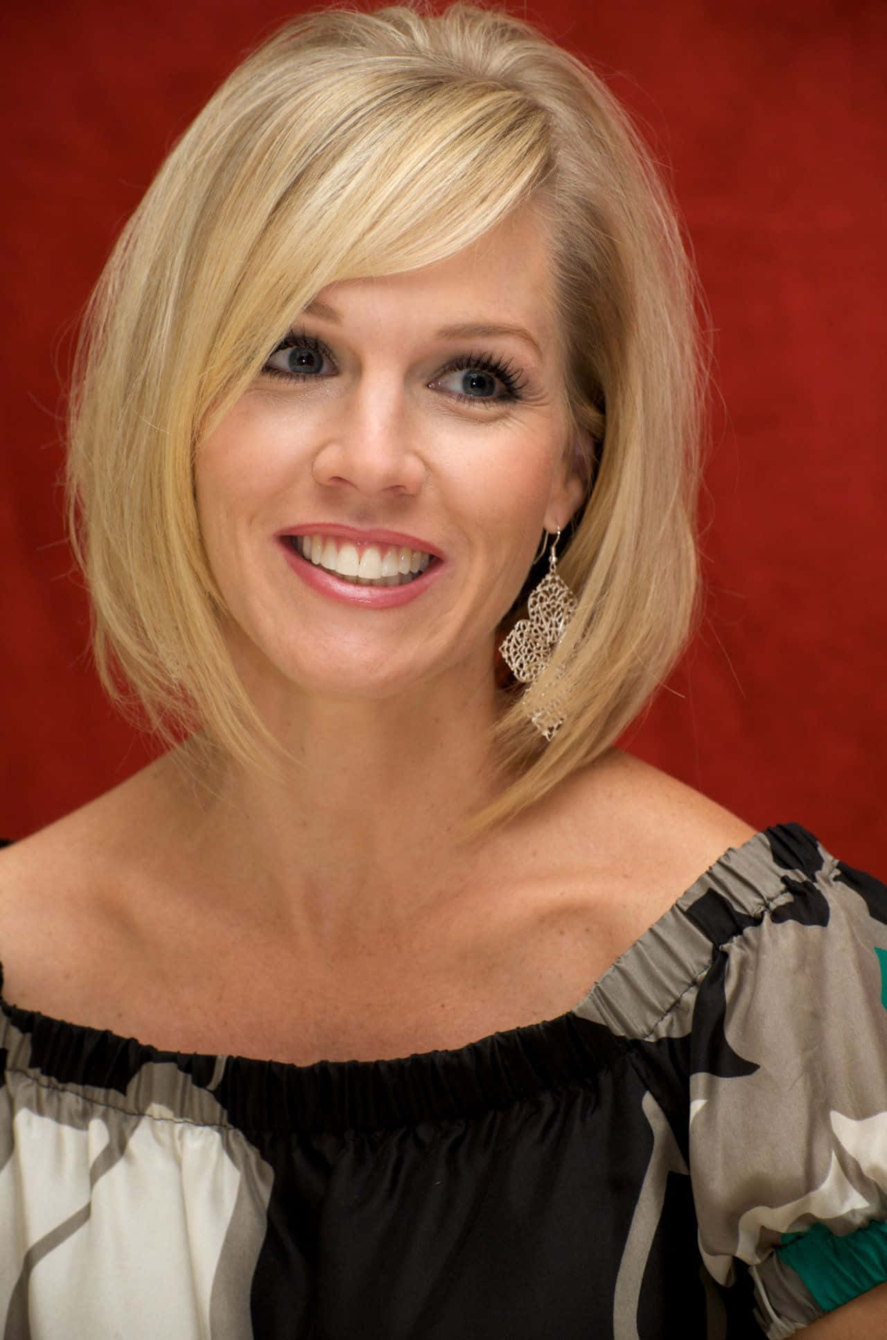 Jennie Garth 桌布 壁紙 — 一位微笑的金髮女士戴著優雅的耳環，在濃郁的紅色背景下穿著時尚的露肩上衣擺姿勢。