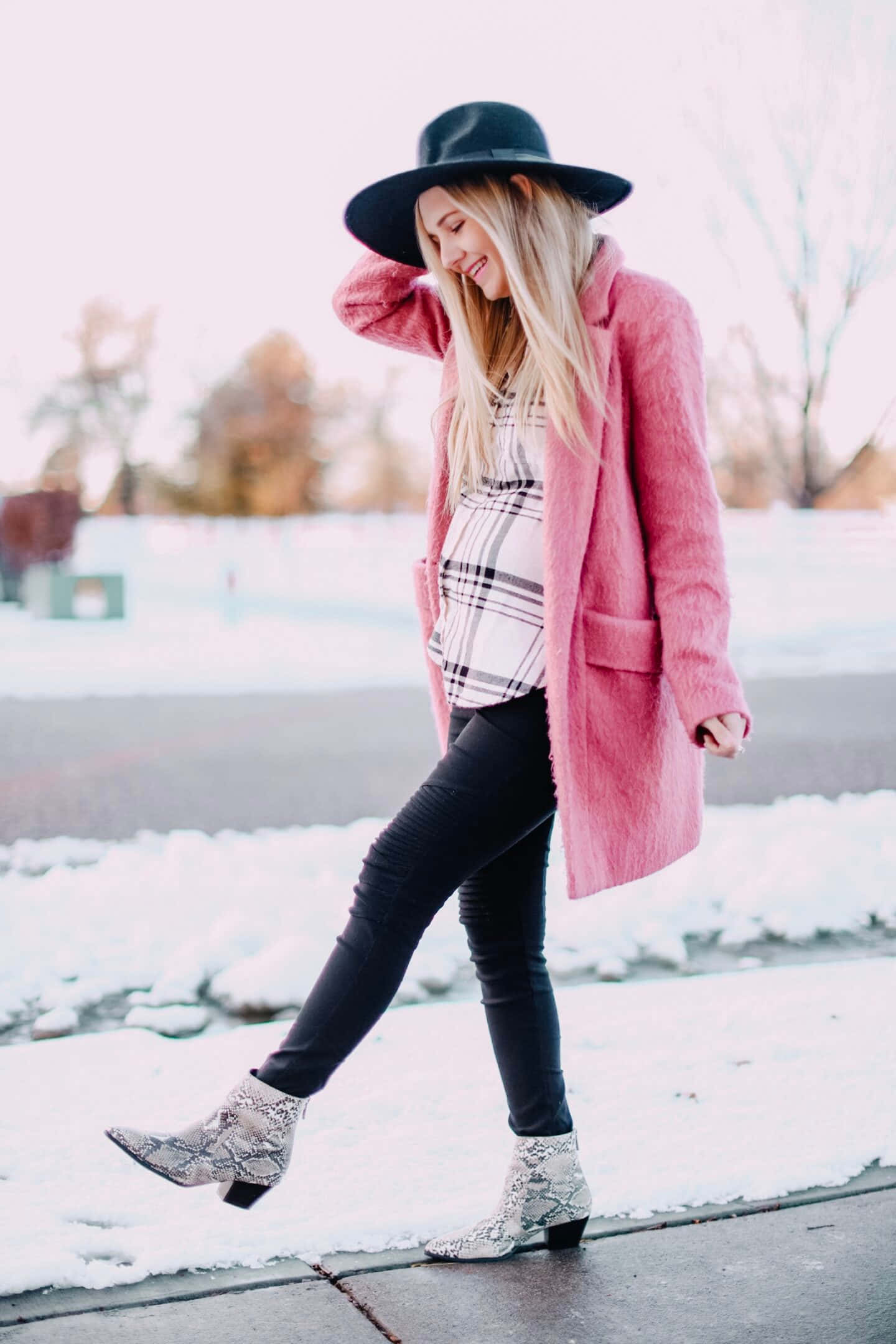 Pink Coat 桌布 壁紙 — 一位微笑的金髮女子，身穿粉紅色外套和黑色帽子，走過白雪皚皚的冬季景色。