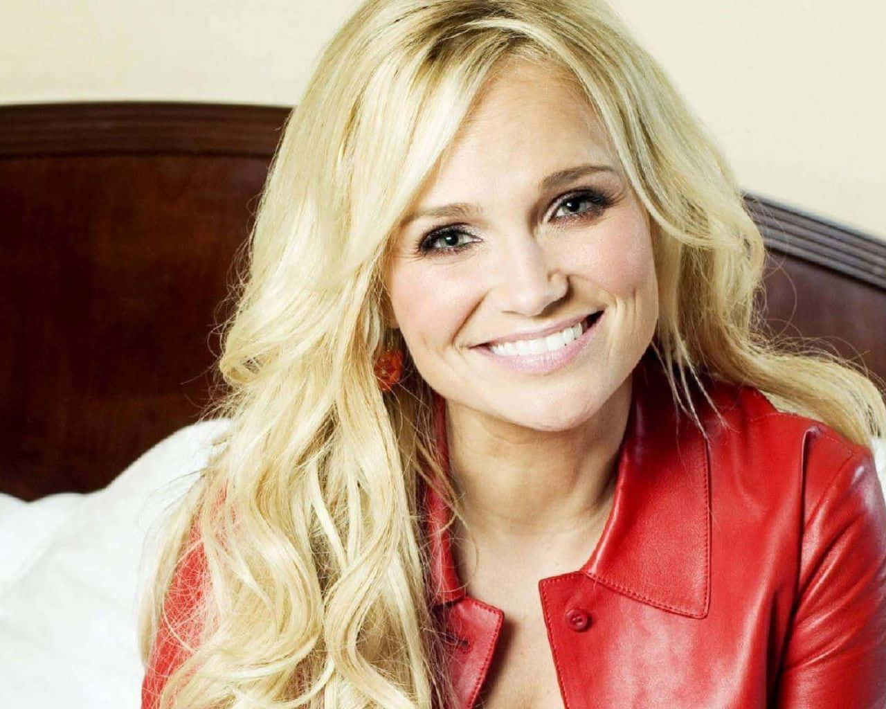 kristin chenoweth 桌布 壁紙 — 一位微笑的金髮女子穿著紅色皮褸，坐在有木製床頭板的床上。
