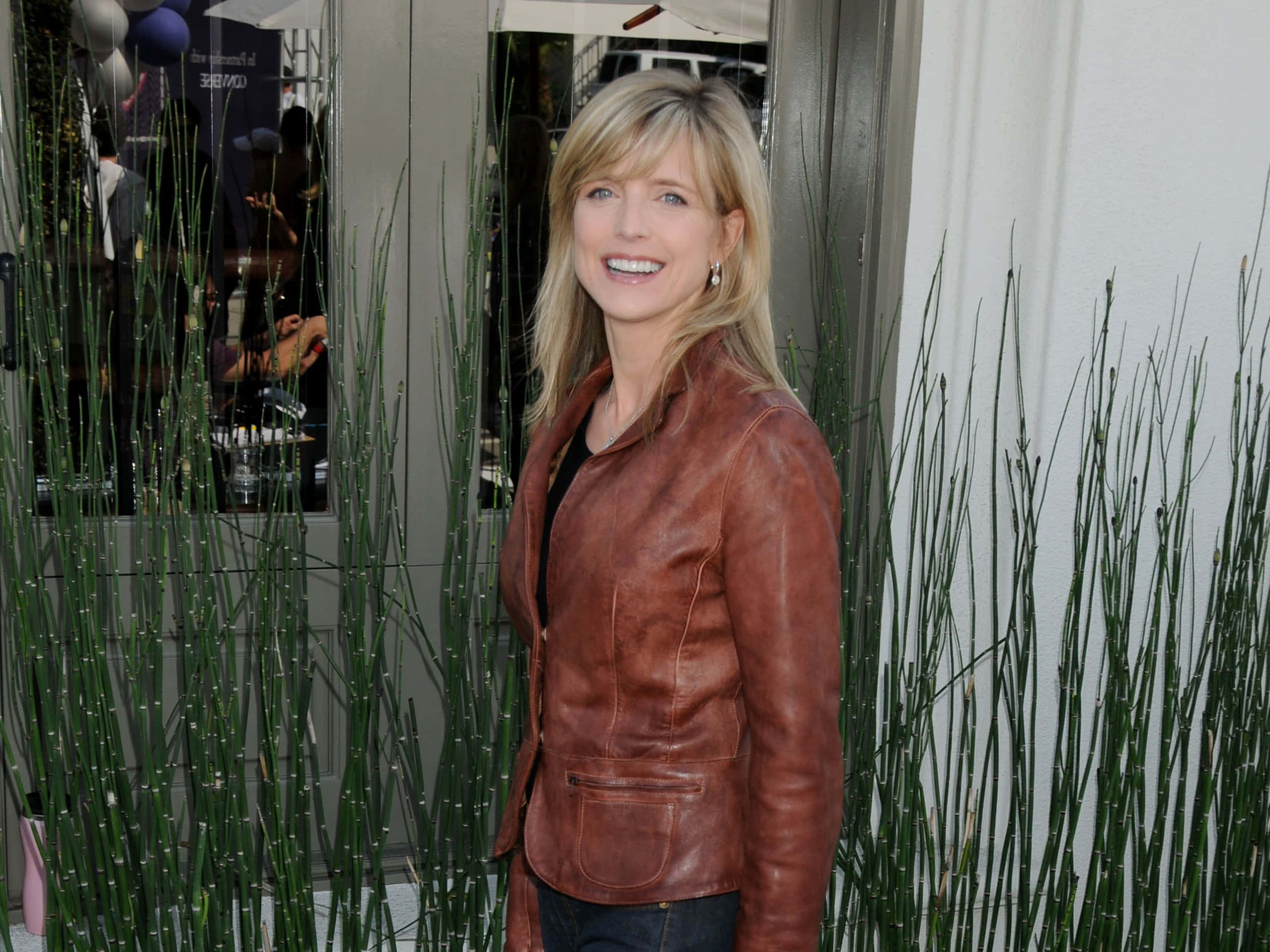 Courtney Thorne Smith 桌布 壁紙 — 一位微笑的金髮女子身穿棕色皮褸，站在玻璃門附近高高的綠色蘆葦旁。