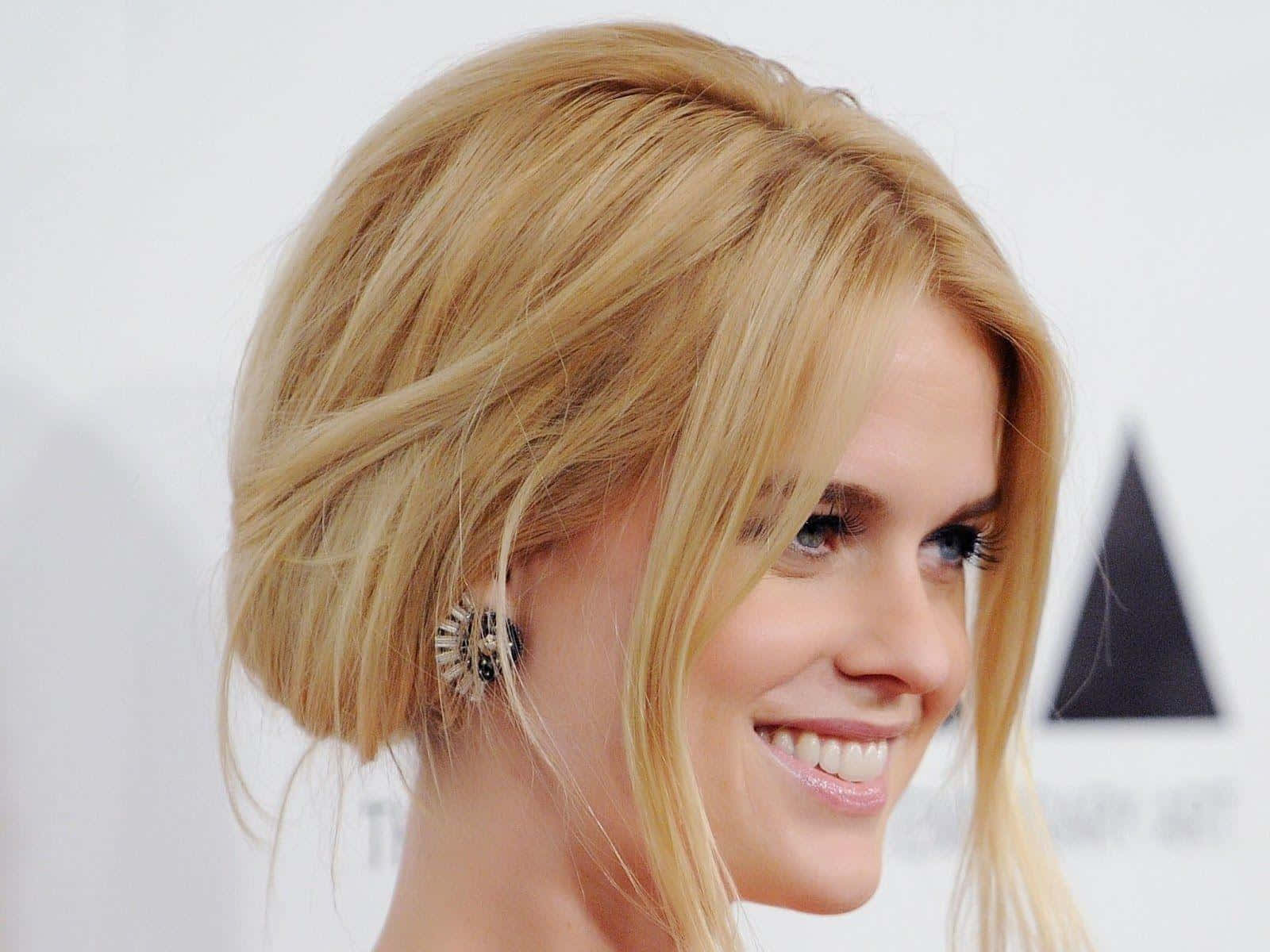 alice eve 桌布 壁紙 — 一位微笑的金髮女性，擁有造型頭髮和華麗耳環，在帶有黑色三角形標誌的白色背景前擺姿勢。