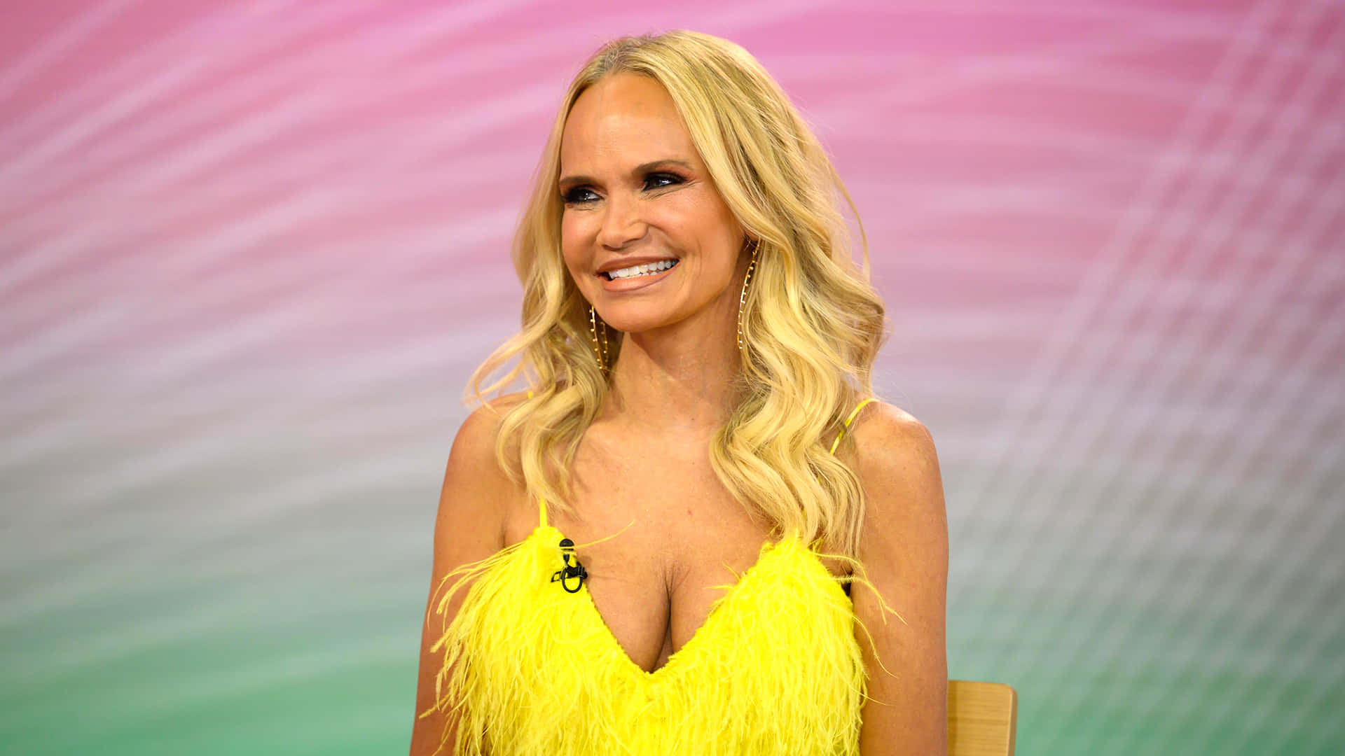 kristin chenoweth 桌布 壁紙 — 一位微笑的金髮女性，身穿亮黃色羽毛上衣，背景是柔和的粉紅和綠色漸變。
