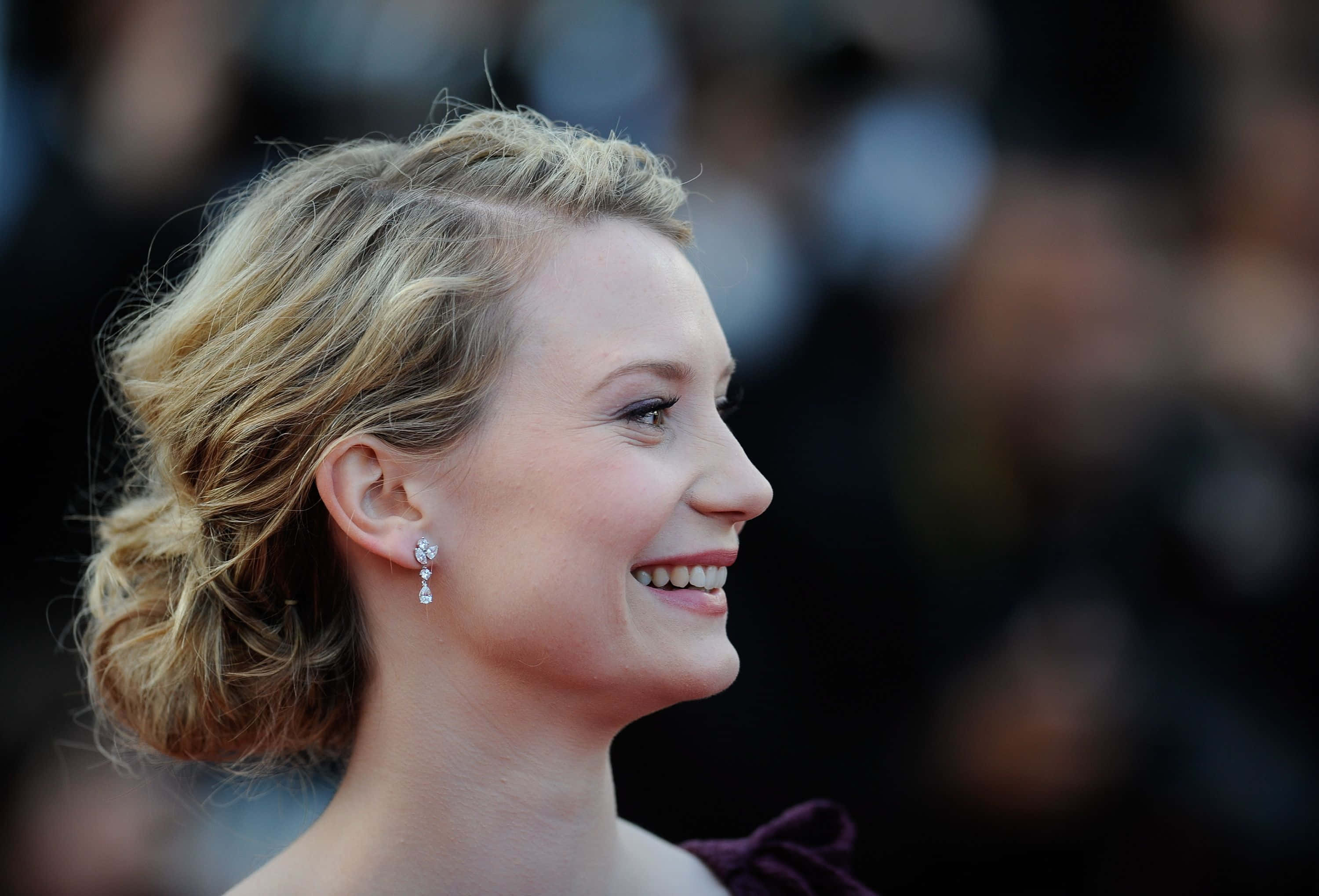 Mia Wasikowska 桌布 壁紙 — 一位微笑的金髮女性，頭髮優雅盤起，戴著閃亮耳環，在華麗活動中看向鏡頭外。
