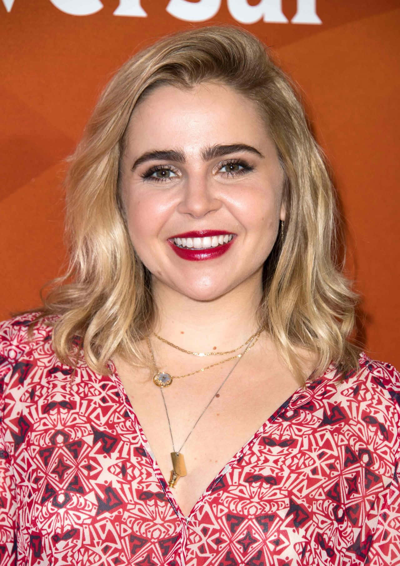 Mae Whitman 桌布 壁紙 — 一位微笑的金髮女性塗著紅色口紅，穿著圖案紅色襯衫，背景為橙色。