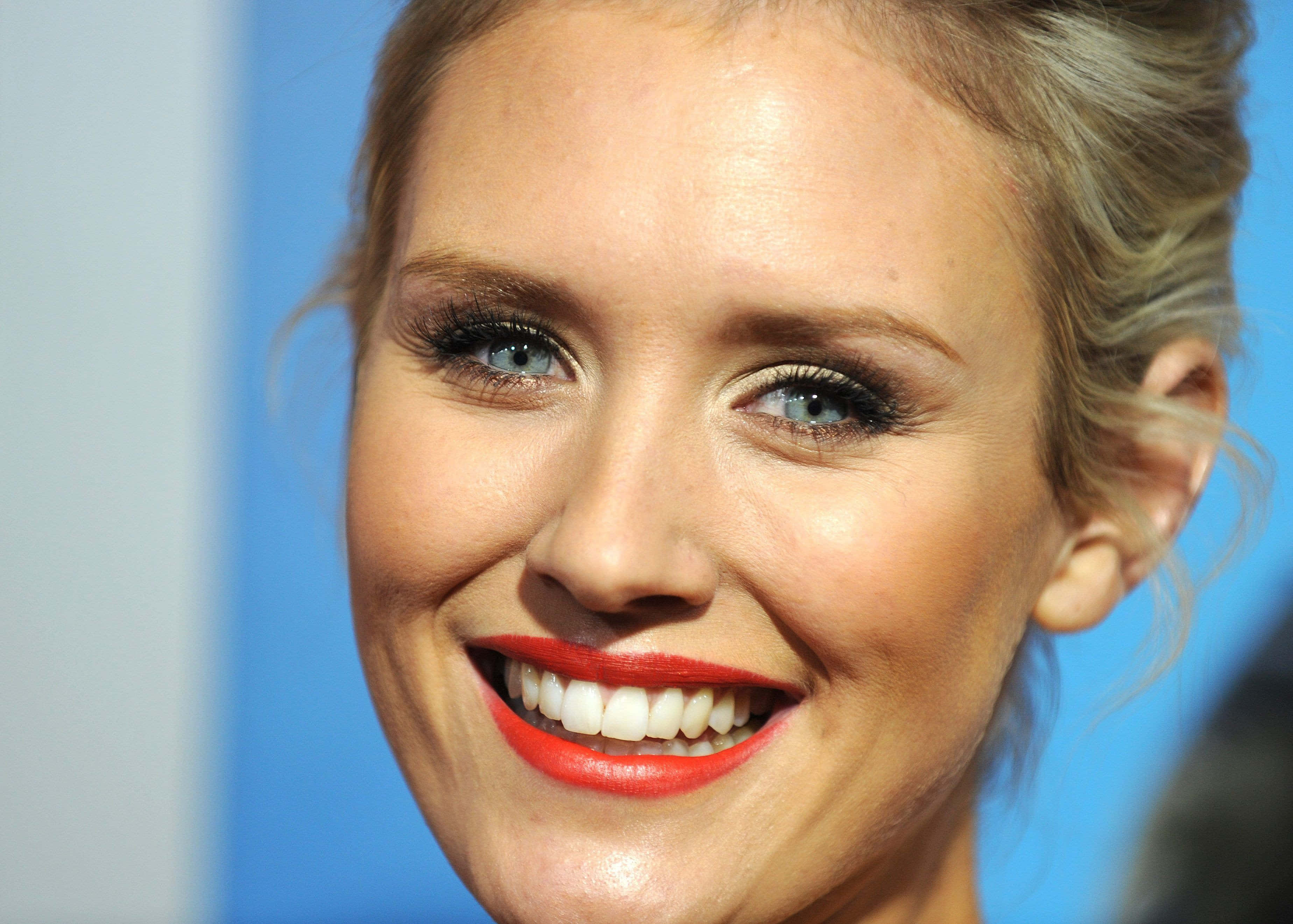 Nicky Whelan 桌布 壁紙 — 一位微笑的金髮女性特寫肖像，擁有明亮的藍色眼睛和鮮豔的紅色唇膏，背景為藍色。