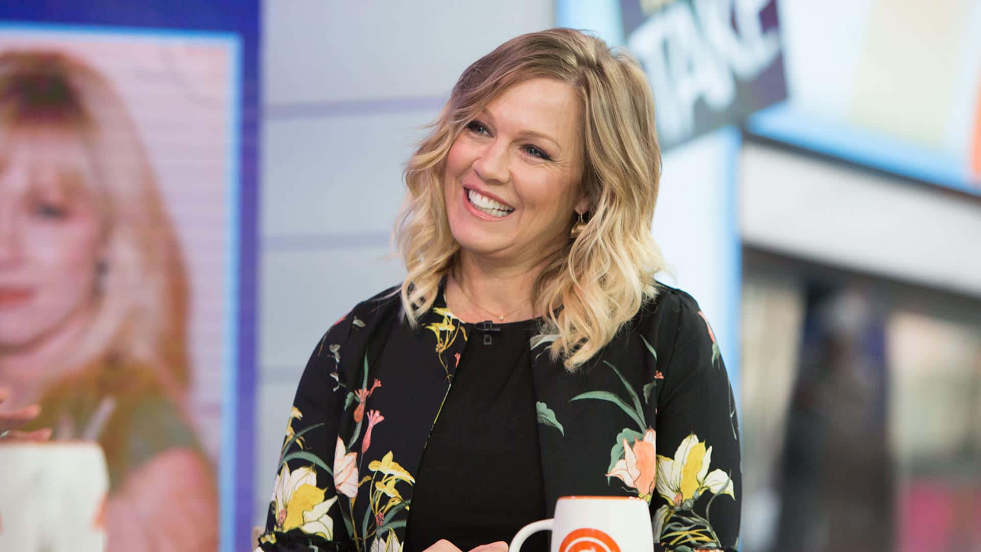 Jennie Garth 桌布 壁紙 — 一位微笑的金髮女性穿著花卉圖案外套，在明亮的脫口秀場景中拿著杯子。