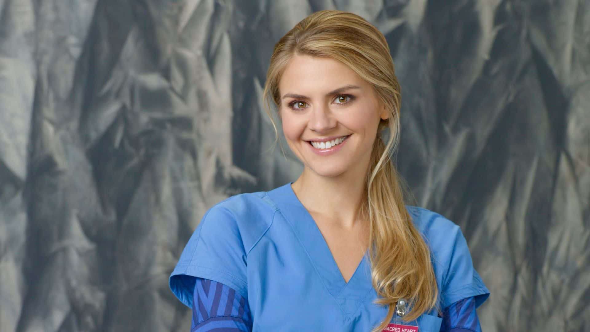 eliza coupe 桌布 壁紙 — 一位微笑的金髮女性穿著藍色工作服，站在有紋理的灰色背景前，表情友善。