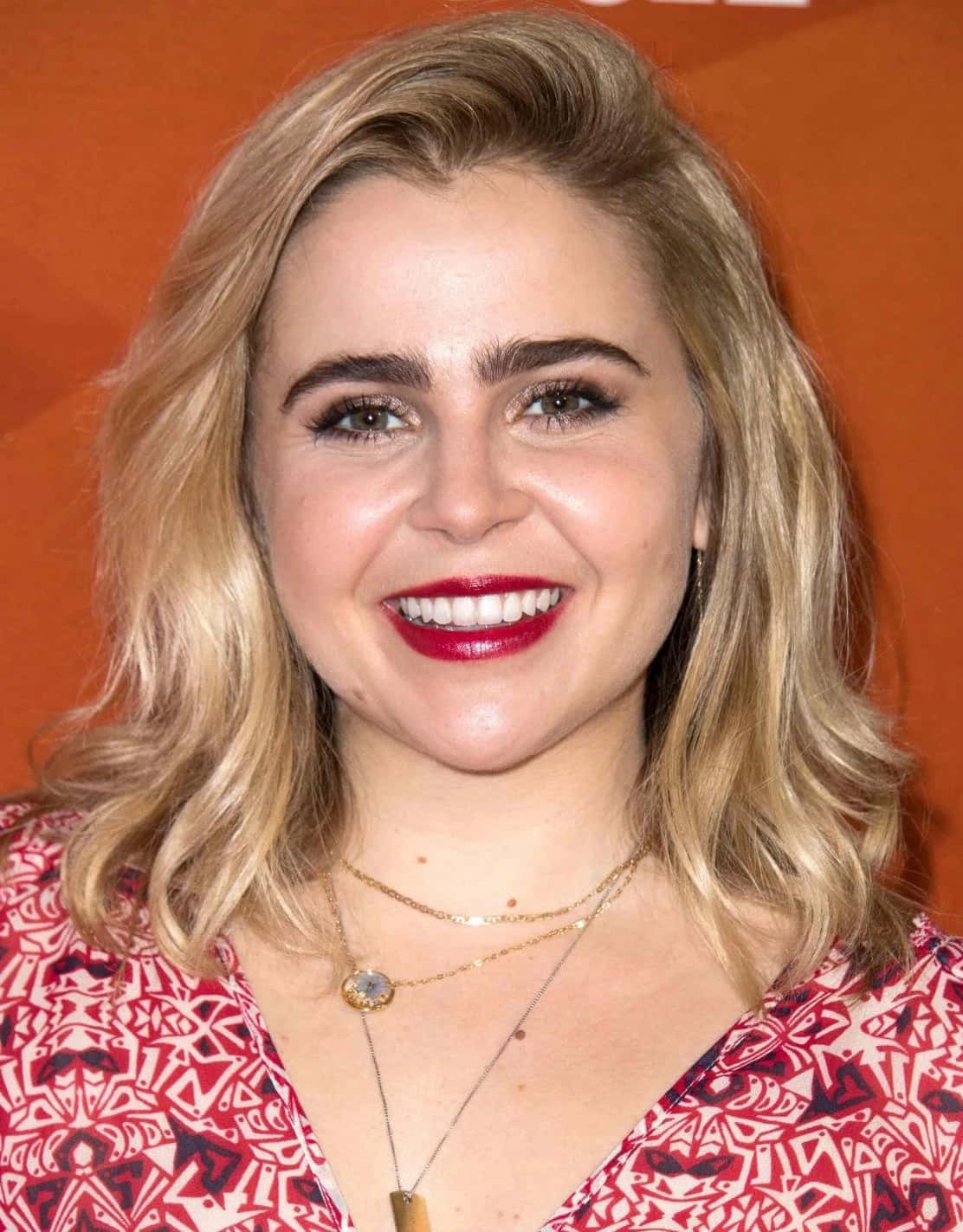 Mae Whitman 桌布 壁紙 — 一位微笑的金髮波浪長髮、塗著大膽紅唇的女性，在溫暖的橙色背景前擺姿勢。