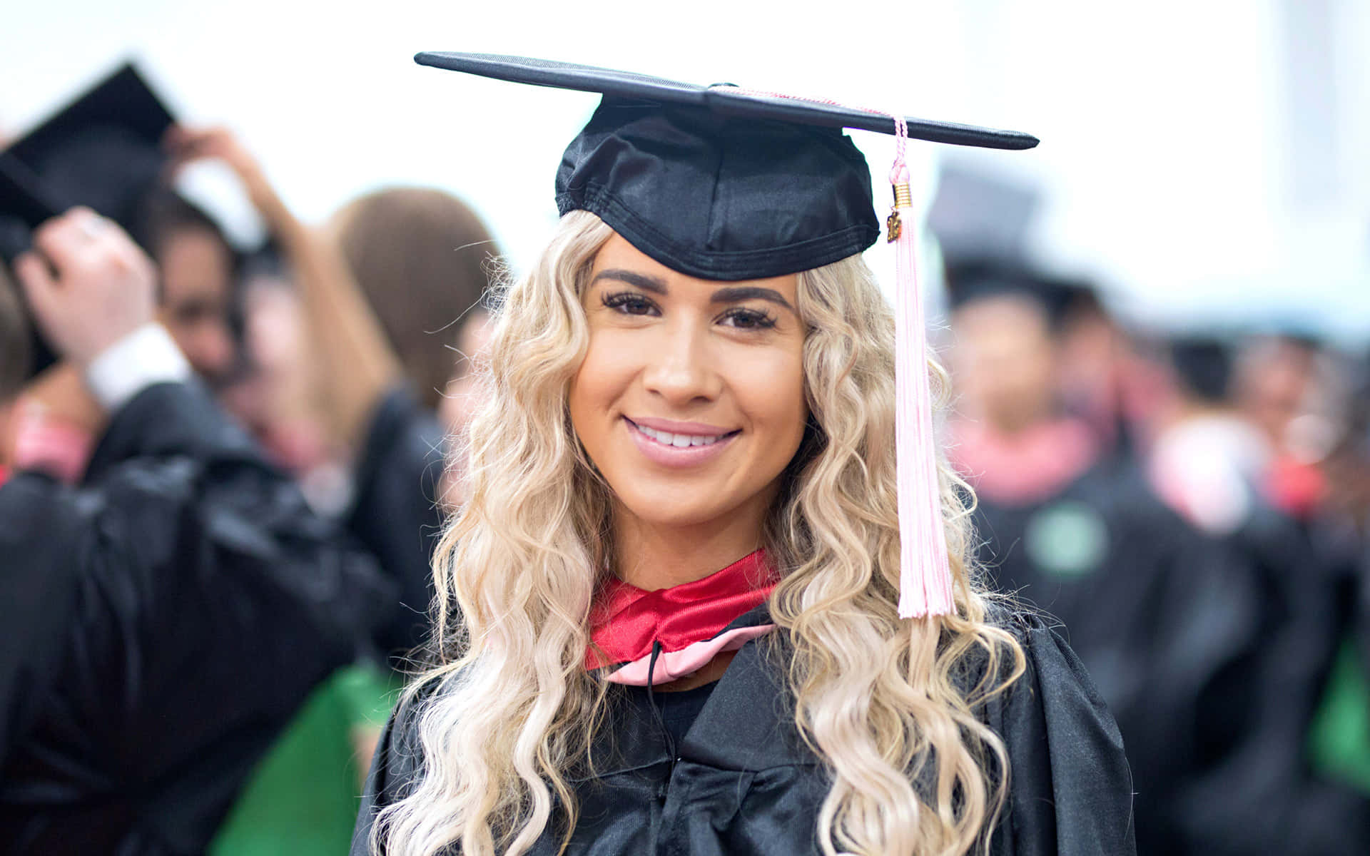 Graduation Ceremony 桌布 壁紙 — 一位微笑的金髮畢業生，身穿黑色學位袍和帽子，帶有粉紅色流蘇，在畢業典禮上模糊的人群中脫穎而出。