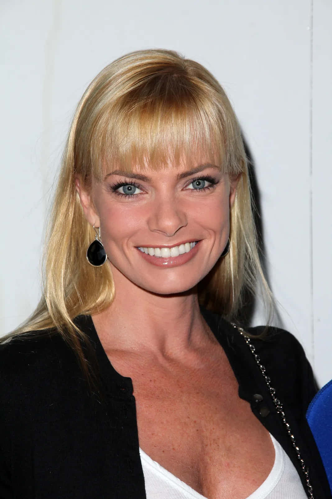 Jaime Pressly 桌布 壁紙 — 一位微笑的金髮藍眼女子，戴著黑色淚滴形耳環，身穿黑色外套內搭白色上衣，背景為純白色。