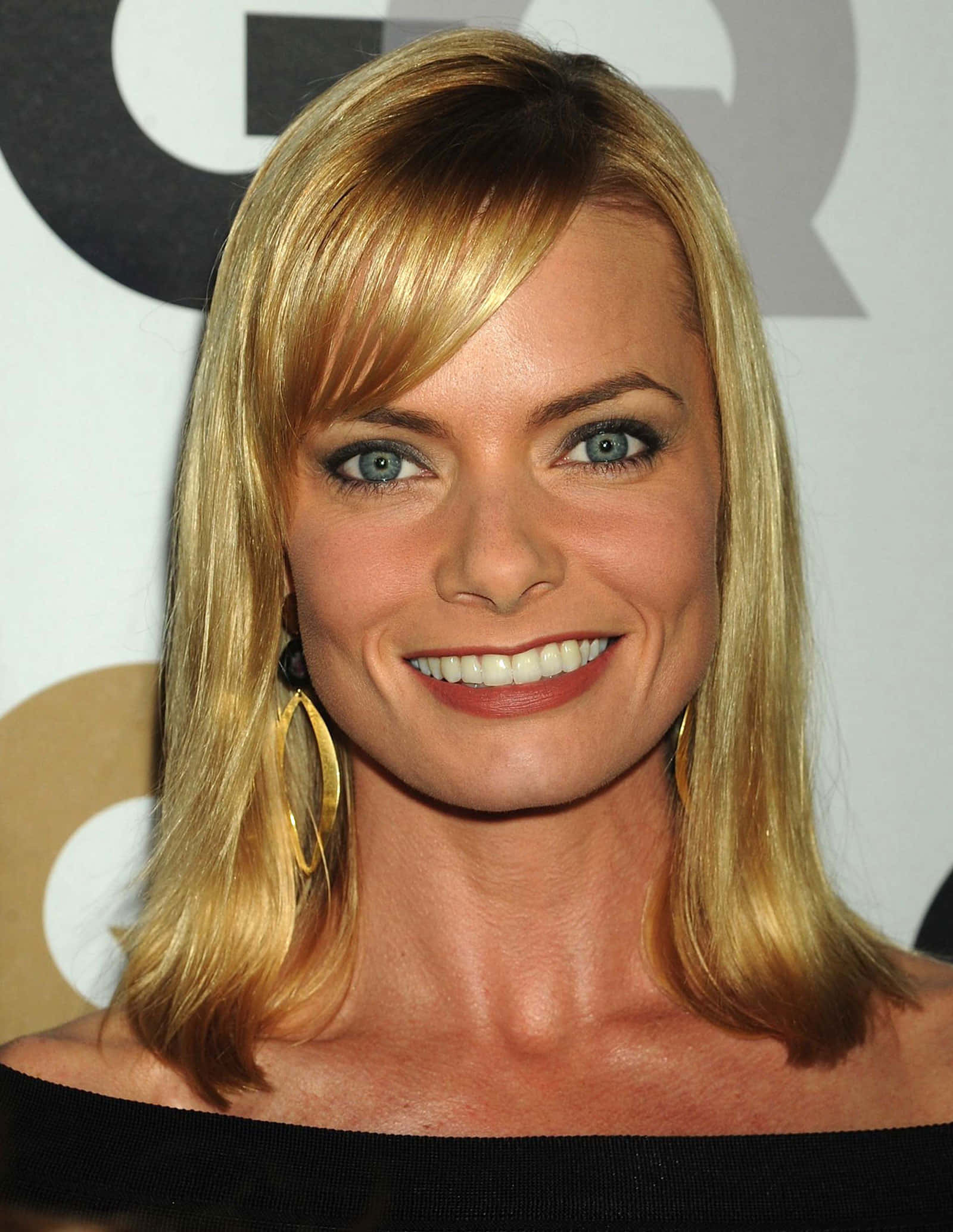 Jaime Pressly 桌布 壁紙 — 一位微笑的金髮藍眼女子戴著金色圓環耳環，在GQ品牌背景前擺姿勢。