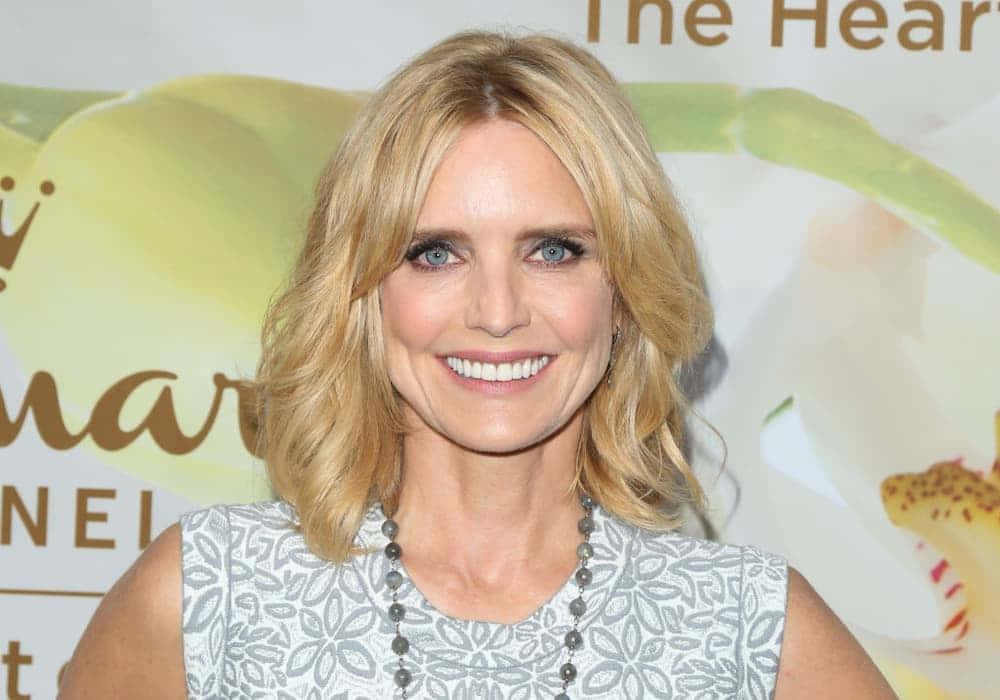 Courtney Thorne Smith 桌布 壁紙 — 一位微笑的金髮藍眼女性，在品牌活動背景前穿著圖案上衣和珠鏈。