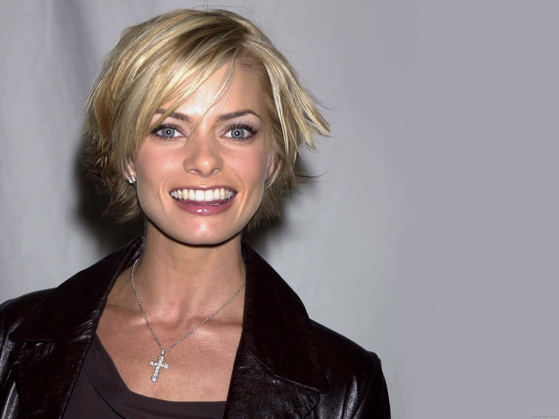 Jaime Pressly 桌布 壁紙 — 一位微笑的金髮藍眼女性，身穿深色皮夾克，戴著十字架項鍊，背景為純灰色。