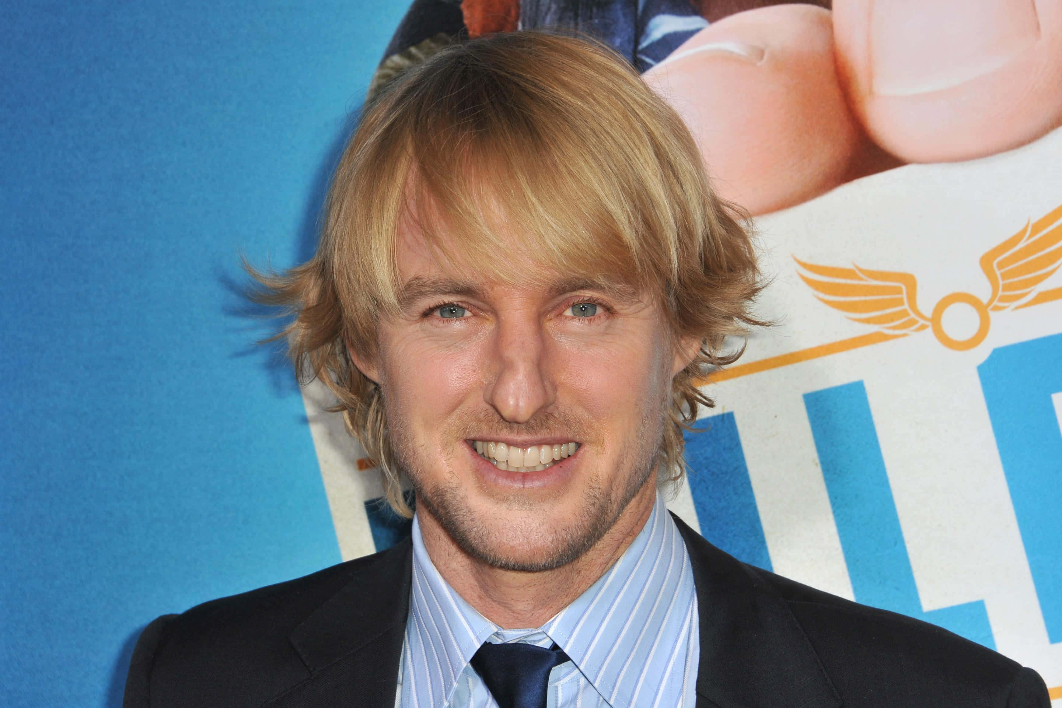 Owen Wilson 桌布 壁紙 — 一位微笑的金髮藍眼男士，身穿深色西裝和條紋襯衫，背景為藍色促銷背景。