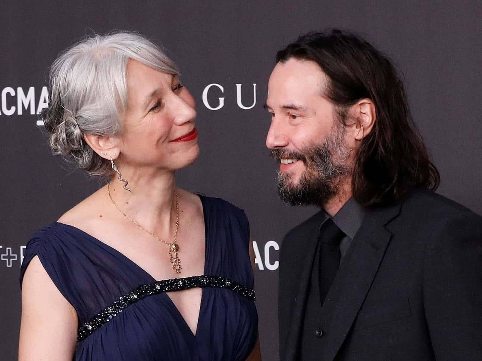 Keanu Reeves 桌布 壁紙 — 一位微笑的銀髮女性和一位長黑髮的男性在品牌背景前於正式活動中分享溫馨時刻。